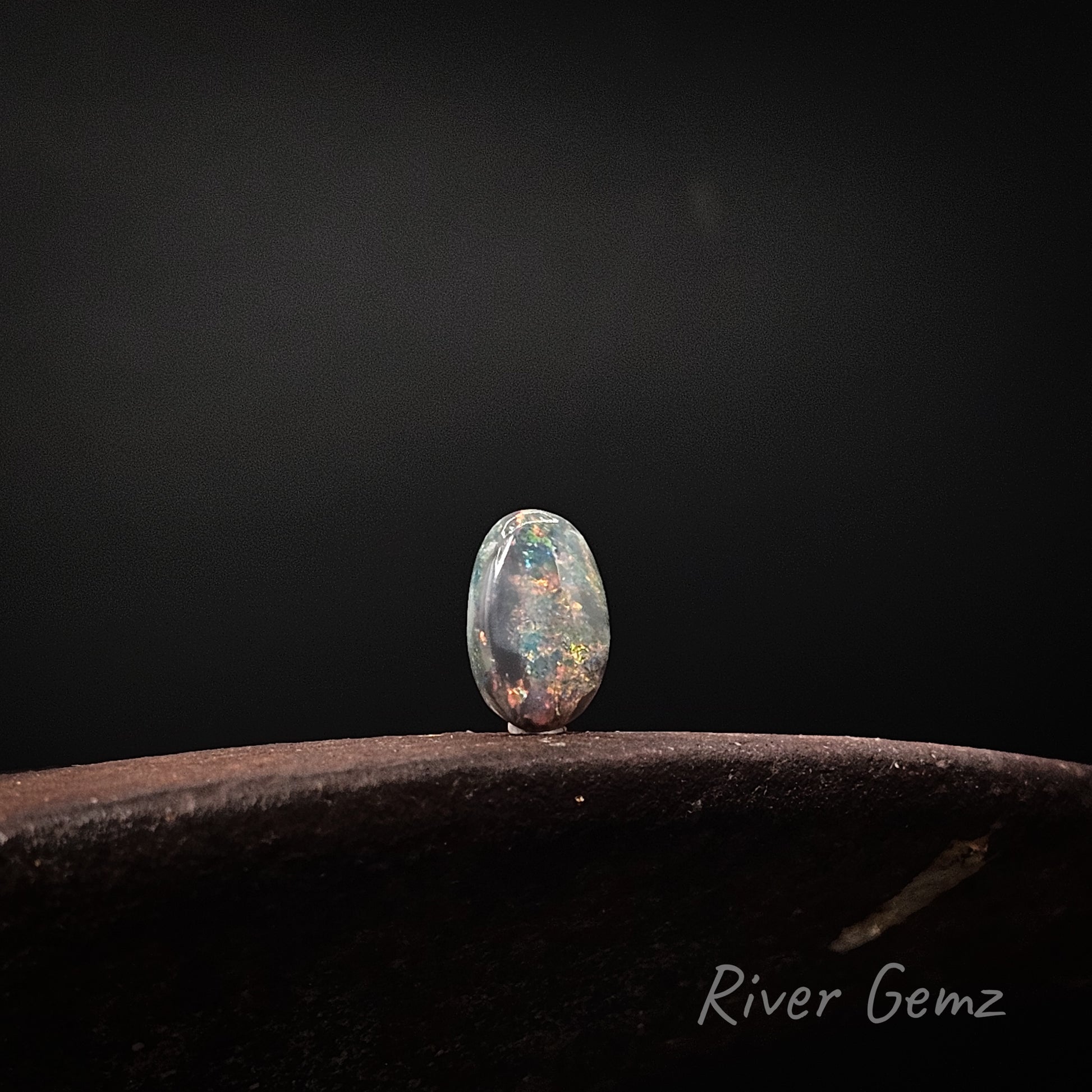 A single oval cabochon gemstone with a vibrant multicolor play-of-colour, standing on a dark textured surface against a dark gray background. The brand name 'River Gemz' is visible in the bottom right corner.