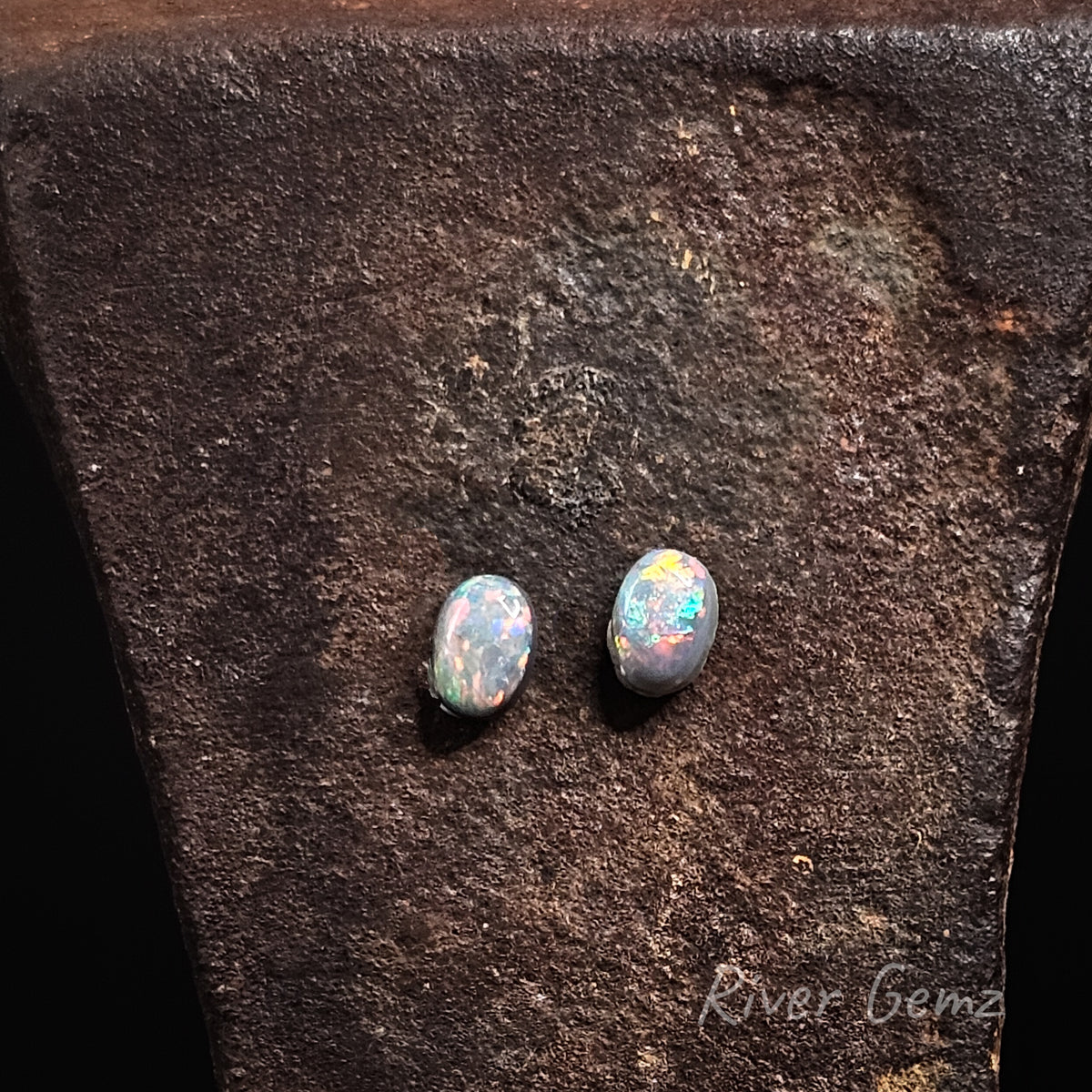 Oval shaped brightly coloured pair of black opals on a rusty coloured background.