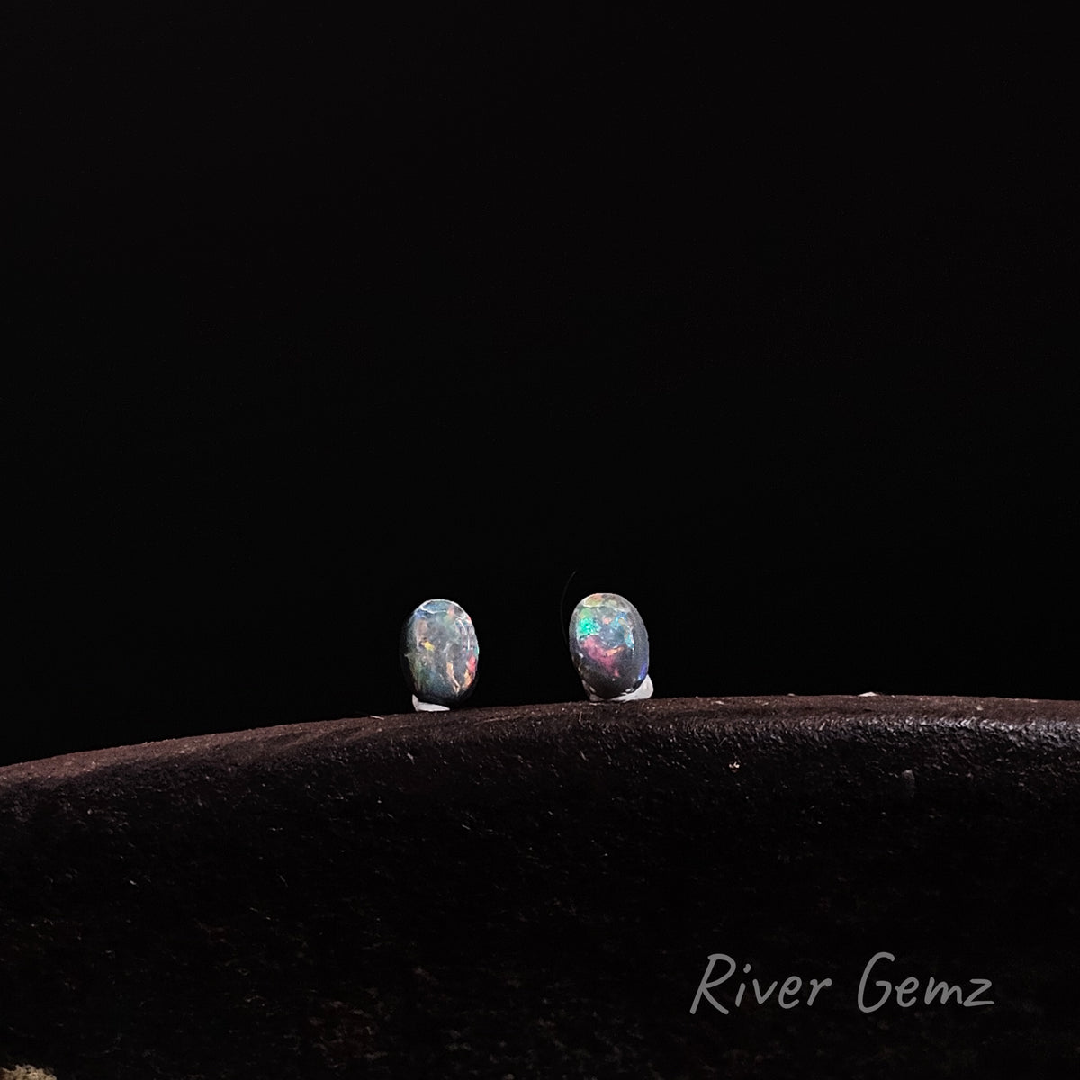 Pair of multi-coloured oval-shaped pair of dark opals standing upright on an antique shoe anvil against a black background.