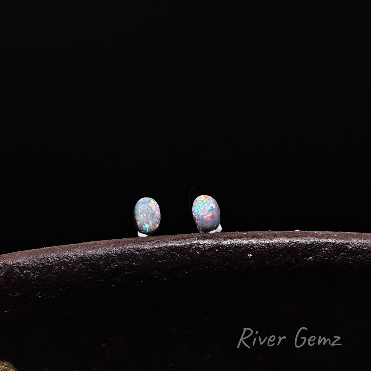 Pair of multi-coloured oval-shaped pair of dark opals standing upright on an antique shoe anvil against a black background with 'River Gemz' watermark in the bottom right hand corner.