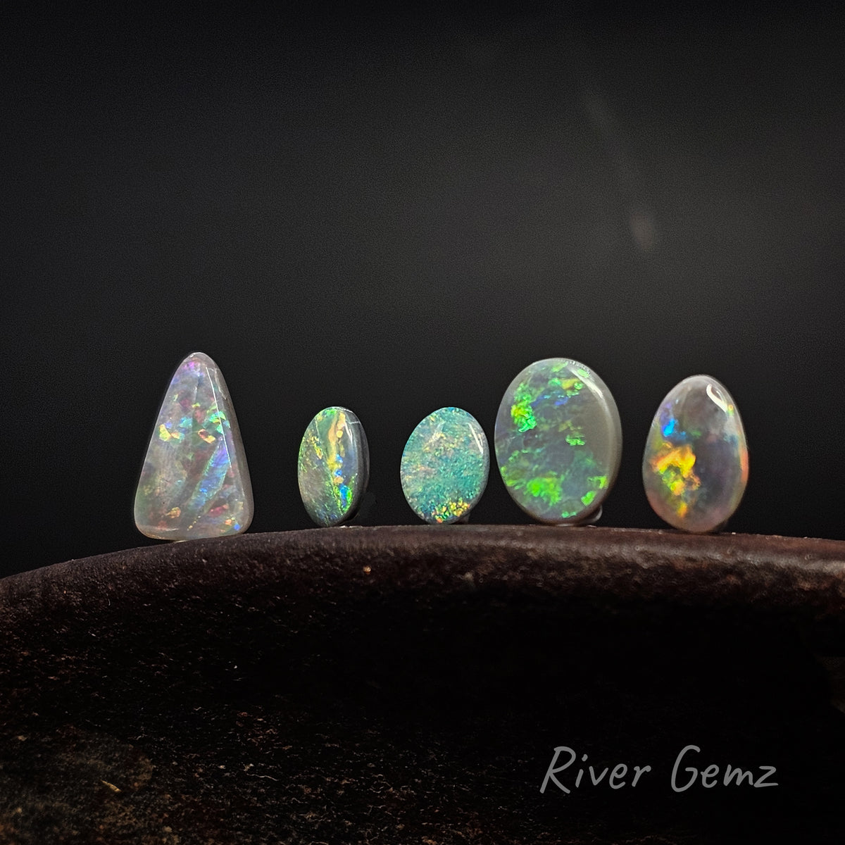5 unset multi-coloured opals standing upright on an shoe anvil with 'River Gemz' watermark in the bottom right hand corner.