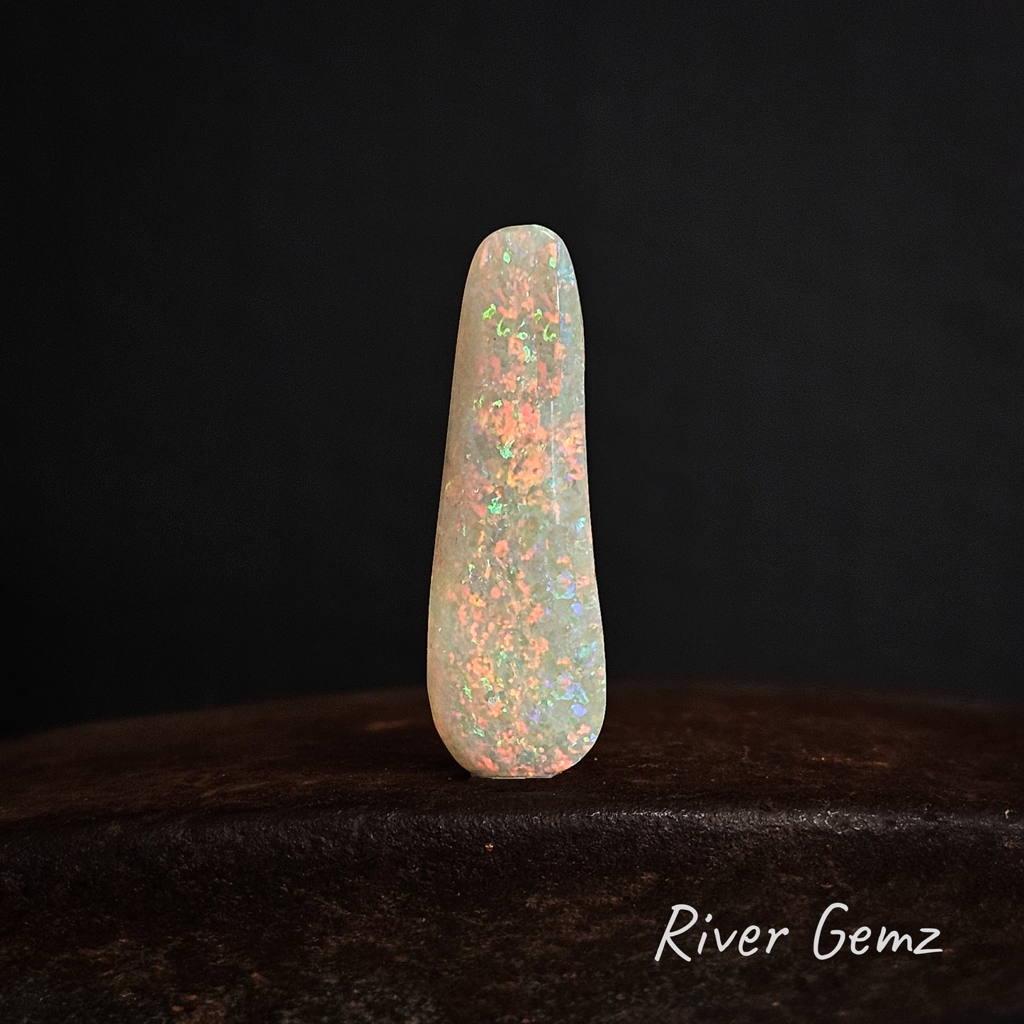 Multi-coloured long white unset opal standing upright on a dark brown surface.