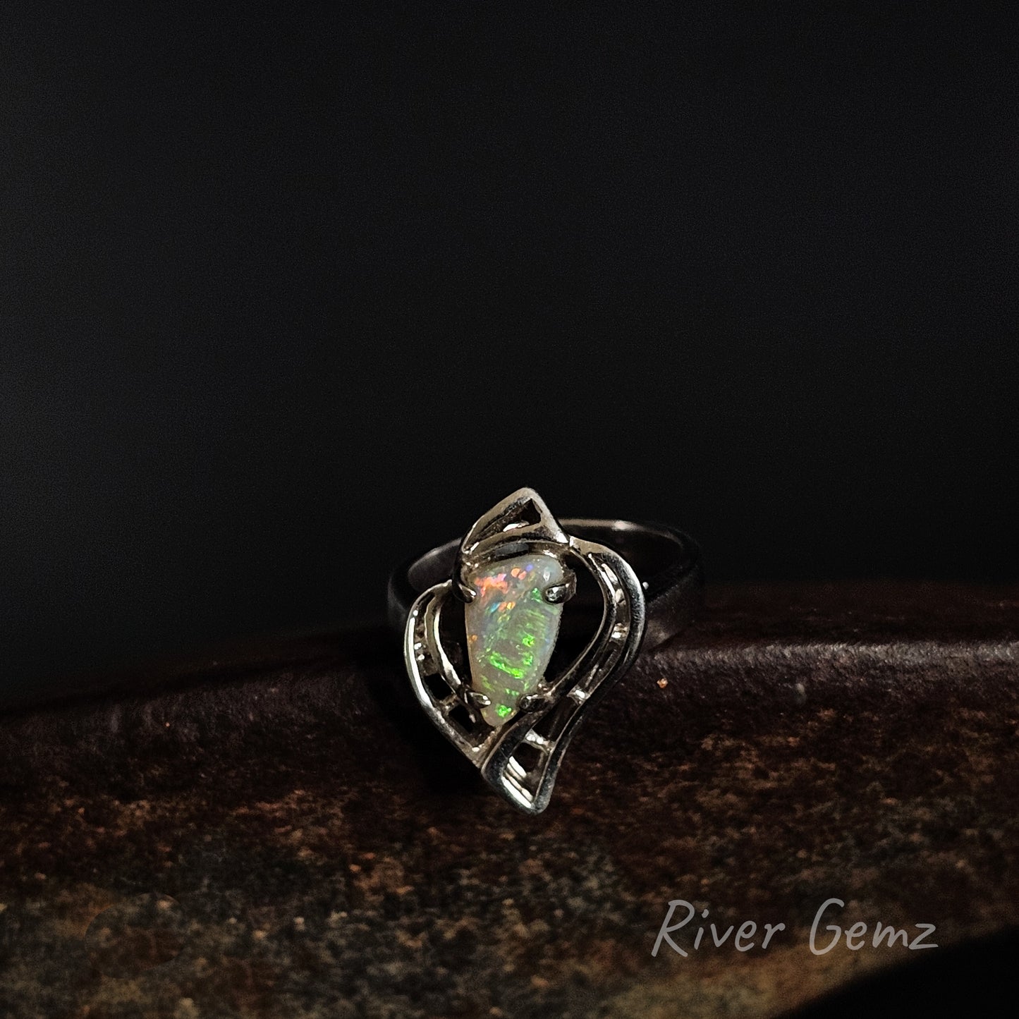 A silver ring featuring a heart-shaped bezel set with a green and orange opal gemstone, resting on a dark textured surface against a black background. The brand name 'River Gemz' is visible in the bottom right corner.