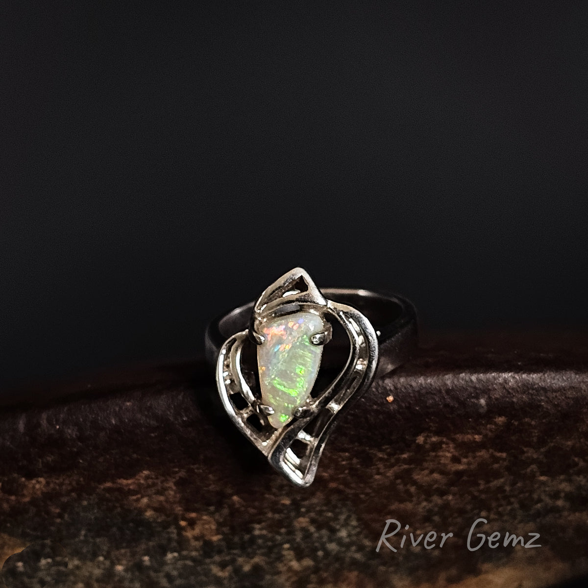 A sterling silver ring featuring a heart-shaped opal gemstone with iridescent green and blue flashes, displayed on a dark textured surface against a black background. The 'River Gemz' brand name is visible in the bottom right corner.