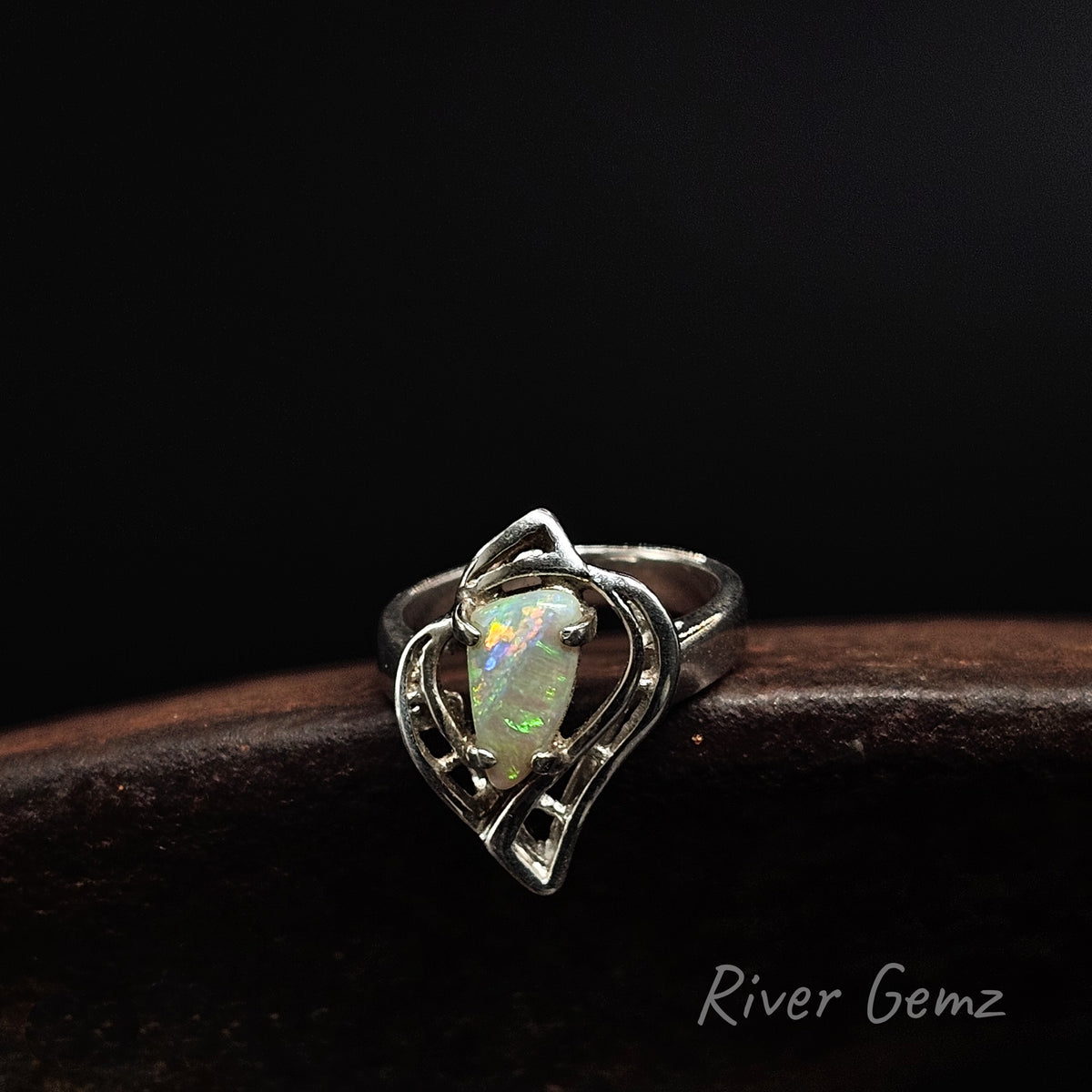 A sterling silver ring featuring a heart-shaped openwork design with a central oval opal gemstone showing vibrant green, orange, and blue flashes, resting on a dark wooden surface against a black background. The 'River Gemz' brand name is visible in the bottom right corner.