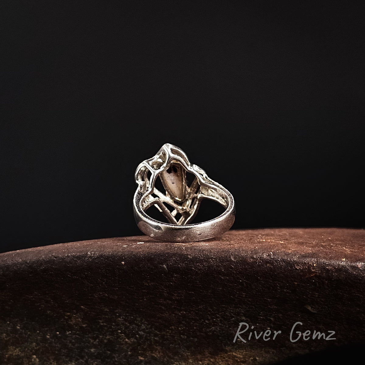 A sterling silver ring with an intricate, openwork filigree design and a central teardrop-shaped stone, resting on a dark brown textured surface against a solid black background. The brand name 'River Gemz' is visible in the bottom right corner.