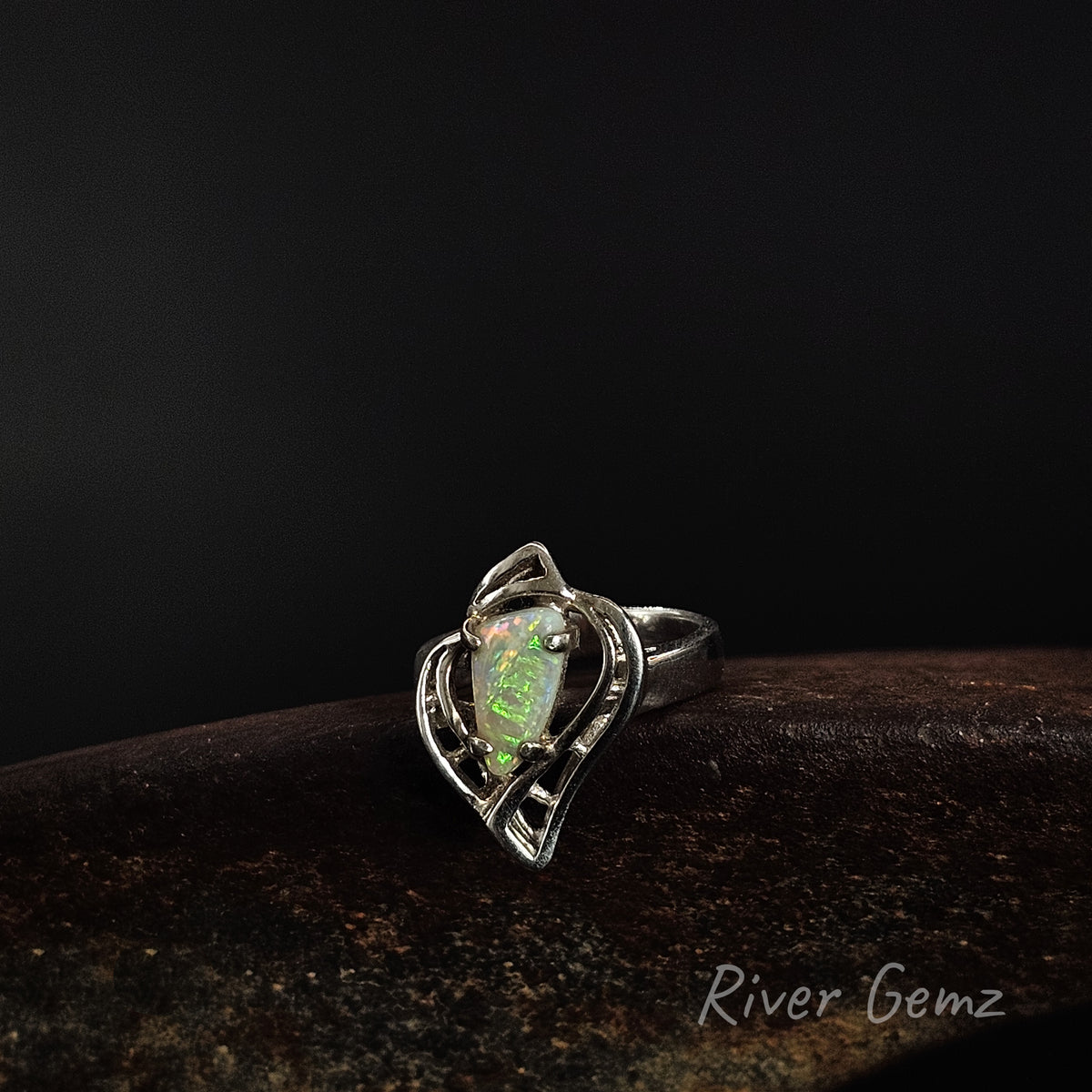 A silver ring featuring a heart-shaped opal gemstone with green and orange flashes, displayed on a dark textured surface against a black background. The brand name 'River Gemz' is visible in the bottom right corner.
