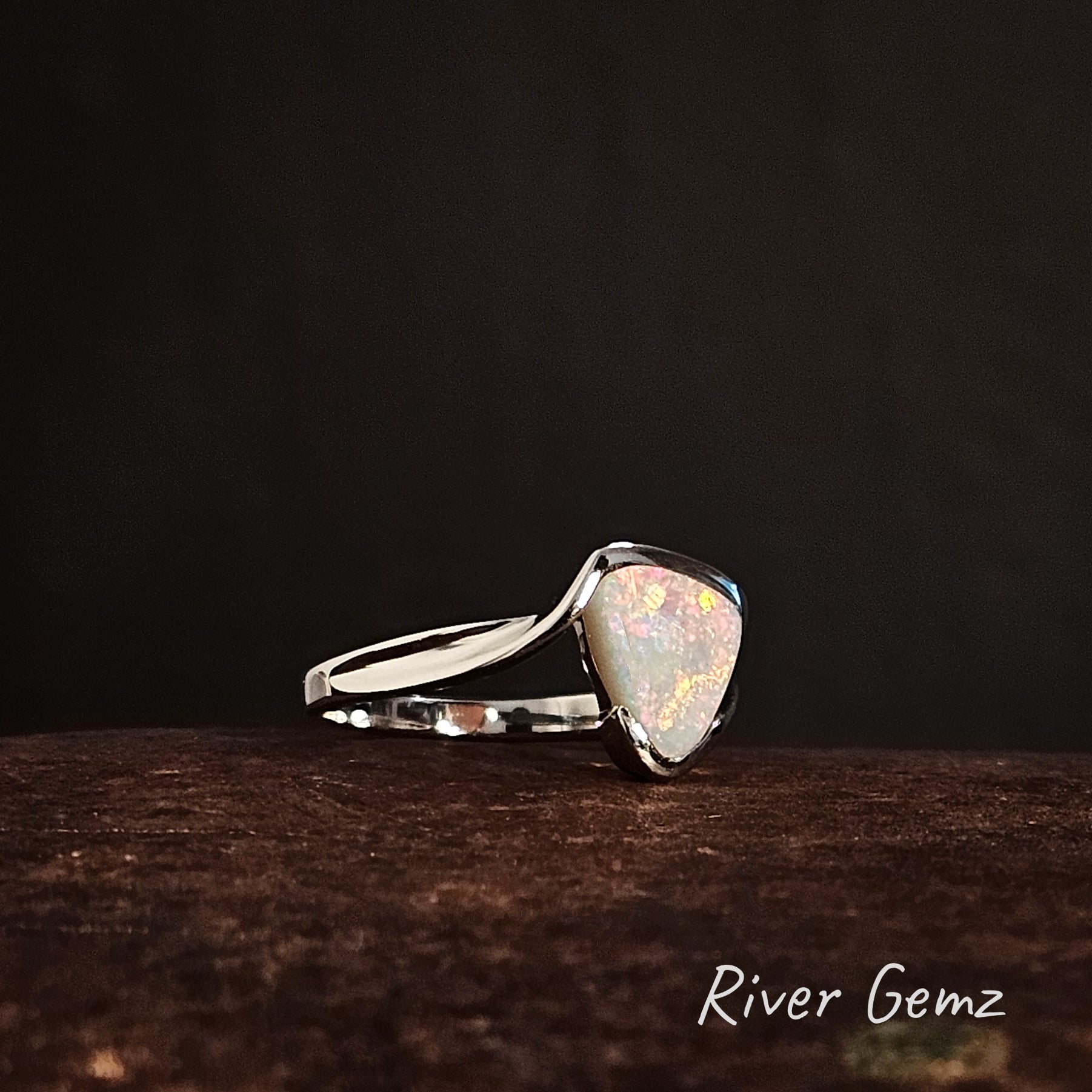 Triangular shaped colourful light opal partial besel set in silver ring on a dark background, branded 'River Gemz'.