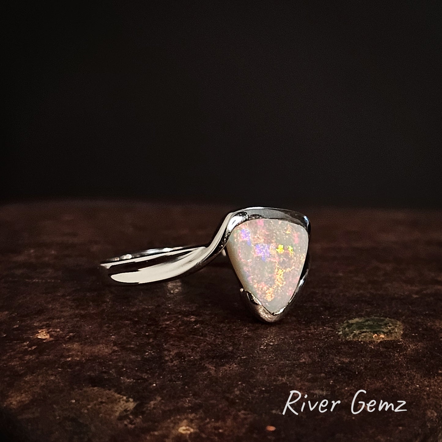 Silver ring with a triangular opal multicoloured stone on a dark surface, branded 'River Gemz'.