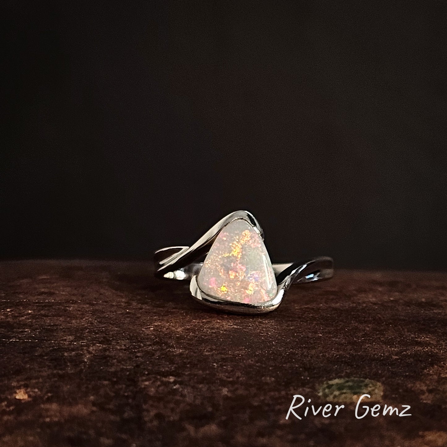 White opal in sterling silver ring on a rust coloured surface with 'River Gemz' branding.