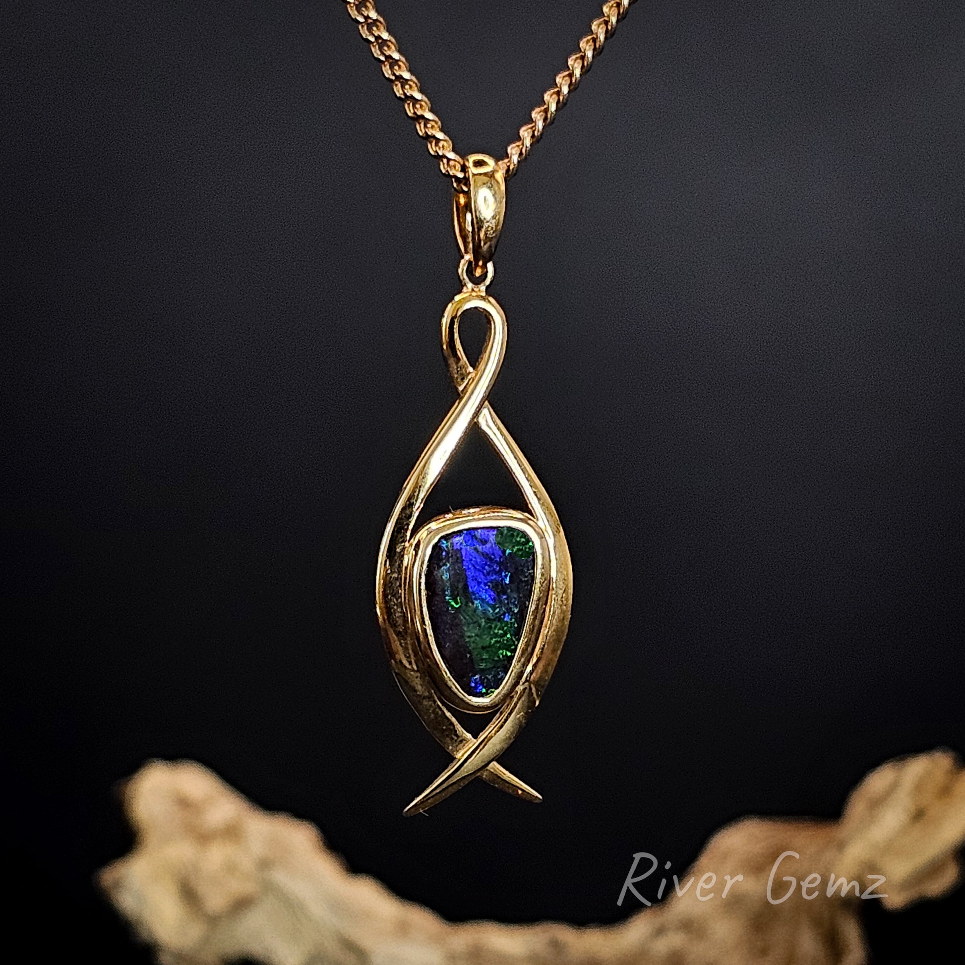 Gold pendant with a colorful gemstone on a dark background, featuring 'River Gemz' branding.