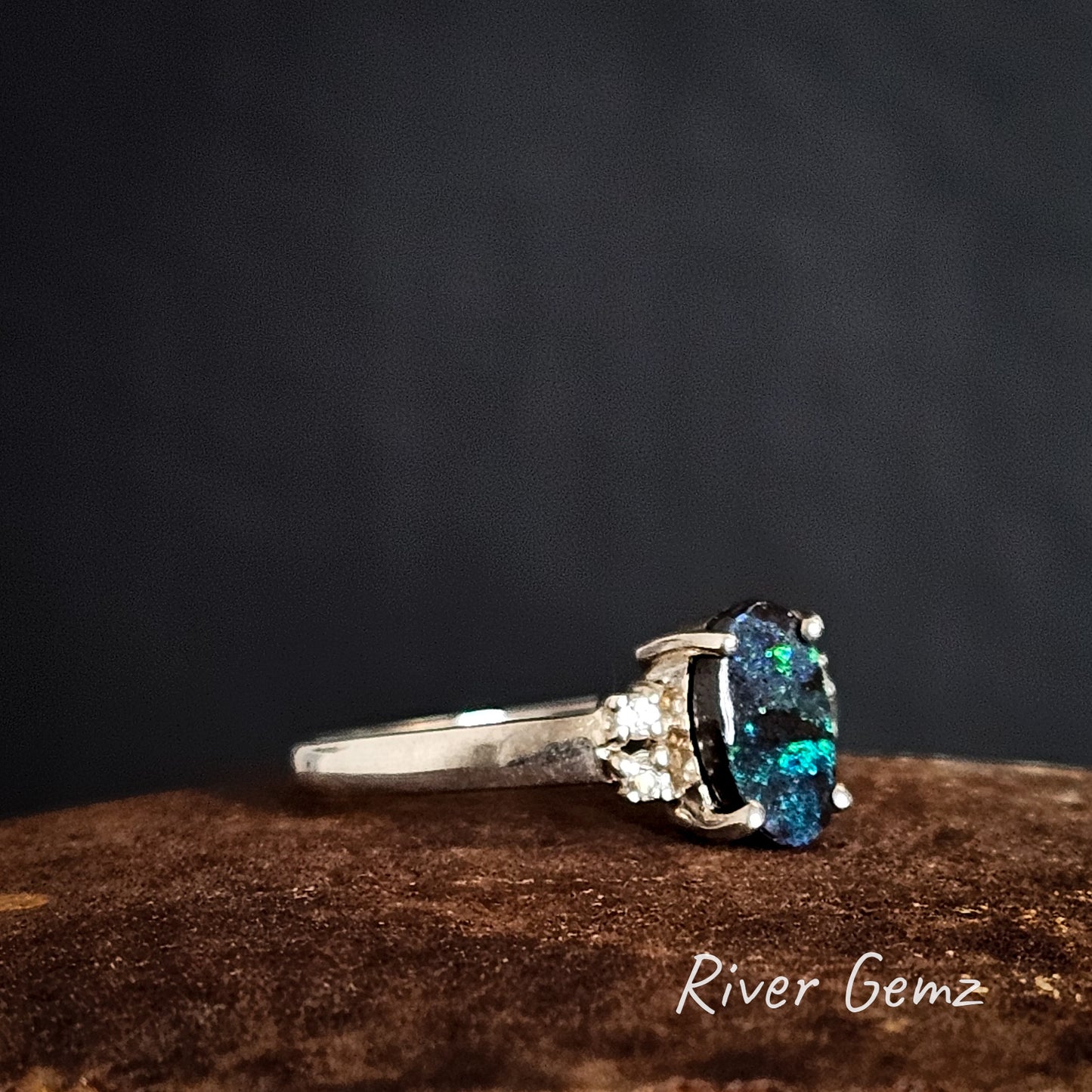 Side view of silver ring with a green and blue boulder opal on a dark background, branded 'River Gemz'.
