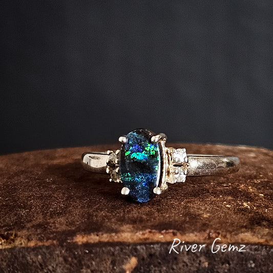 Silver ring with a dark gemstone on a weathered iron shoe anvil, branded 'River Gemz'.