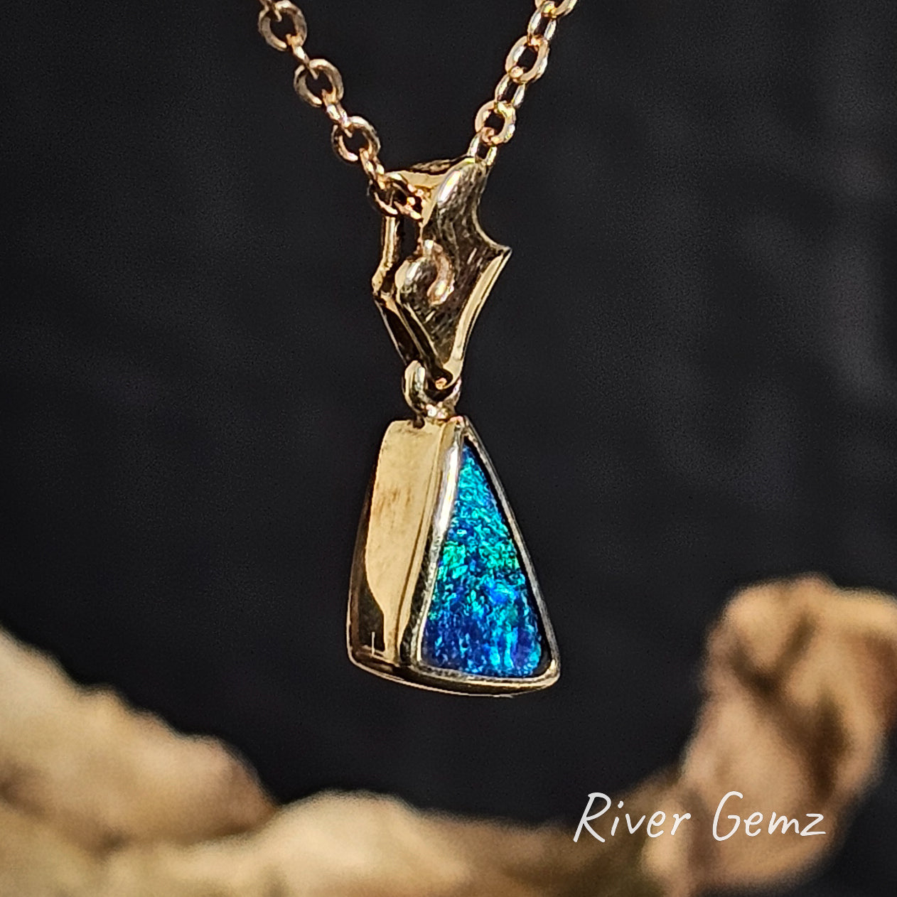Gold pendant with triangular shaped blue/green boulder opal on a chain against a dark background branded 'River Gemz'.