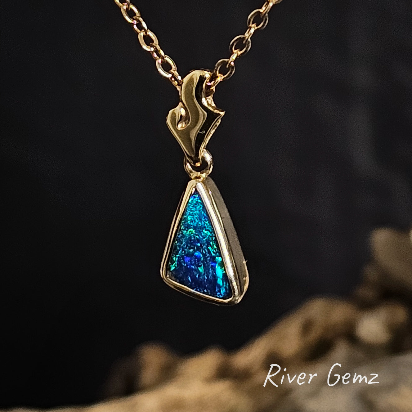 A blue triangular dark opal besel set in yellow gold necklace with a flame-shaped bail  on a dark background branded 'River Gemz'.