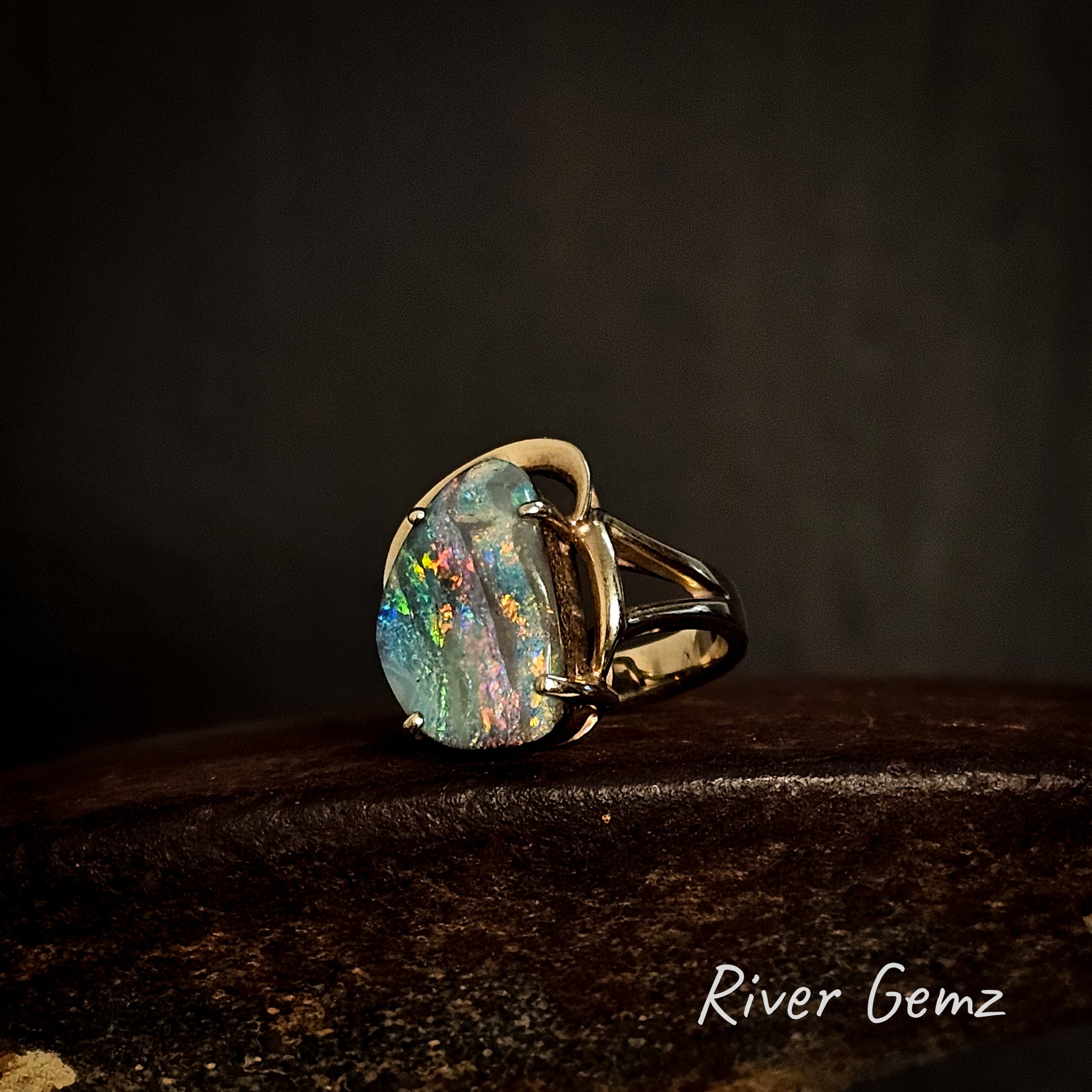 Yellow gold ring with a large colorful boulder opal on a dark background, featuring the brand 'River Gemz'.