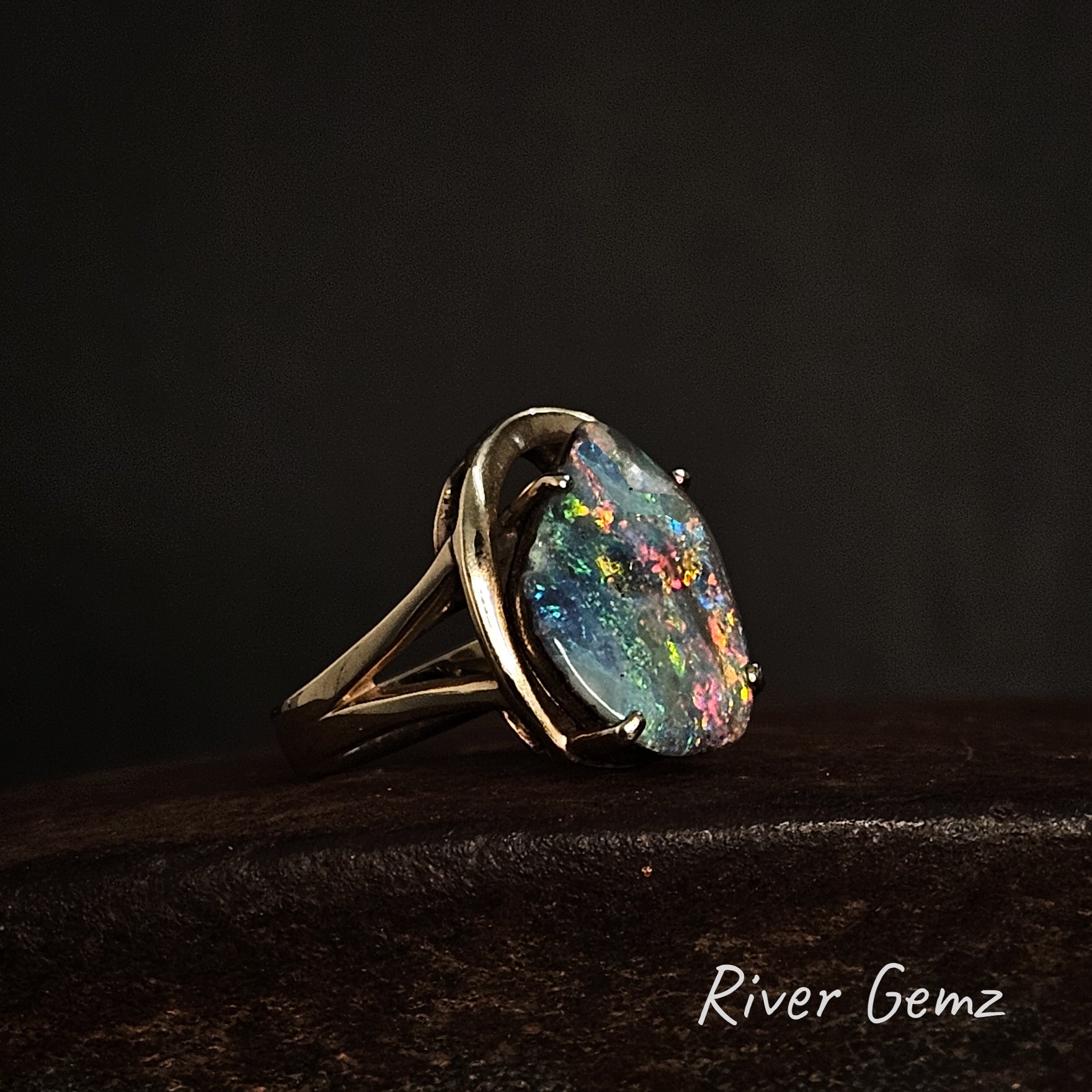 Ring with a free-form cut multi-coloured boulder opal on a dark background, branded 'River Gemz'.