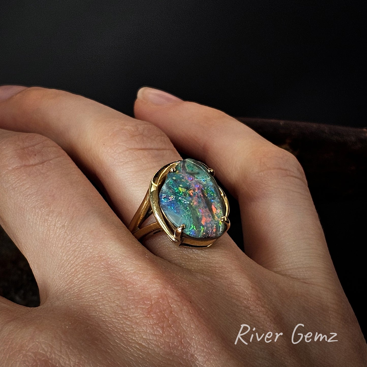 Multi-coloured boulder opal in yellow gold ring on a hand with 'River Gemz' branding.