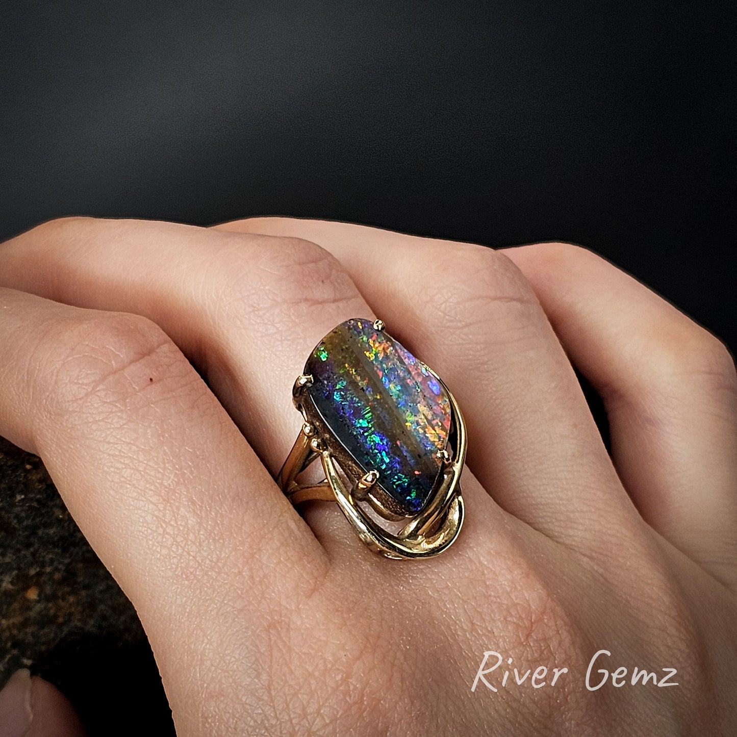 Ring with a colorful gemstone on a hand, branded 'River Gemz'.