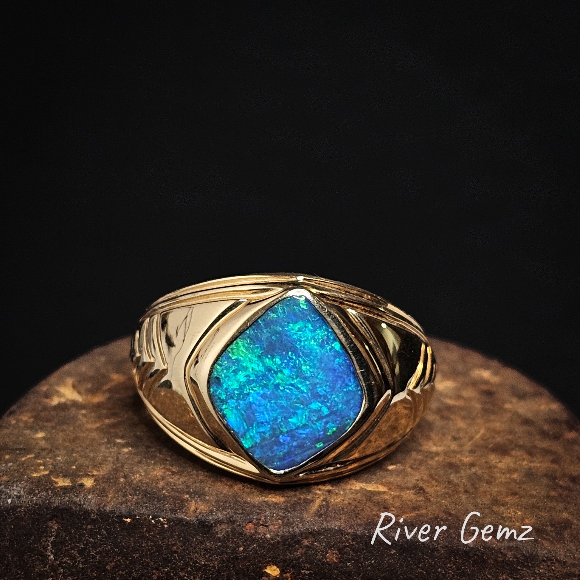 Gold ring with a blue/green diamond shaped boulder opal stone on a dark background, branded 'River Gemz'.