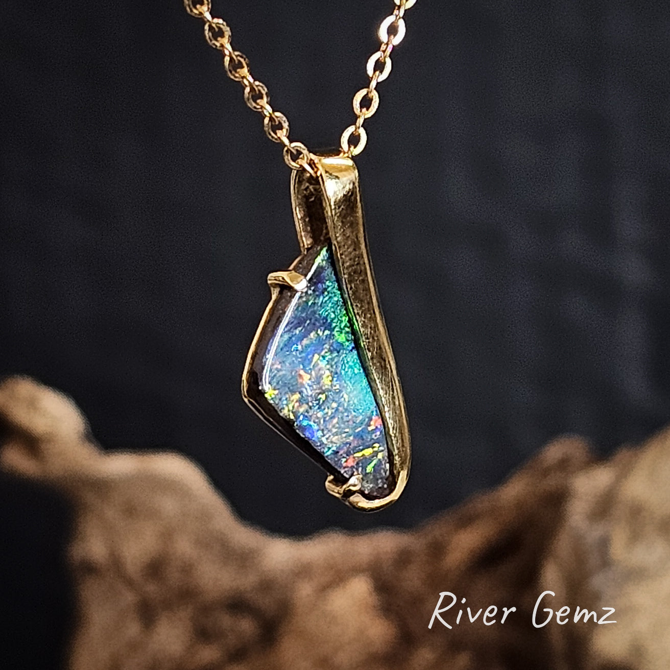 Multi-coloured boulder opal set in yellow gold pendant on a dark background with 'River Gemz' branding.