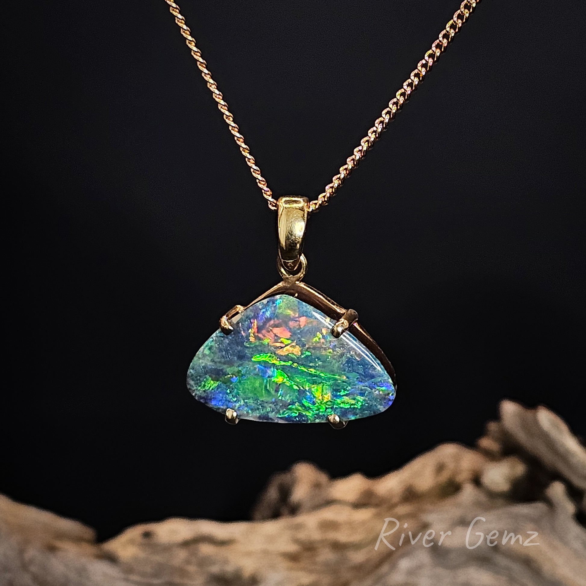 Multi-coloured boulder opal claw set in yellow gold pendant on a gold chain with a dark background.