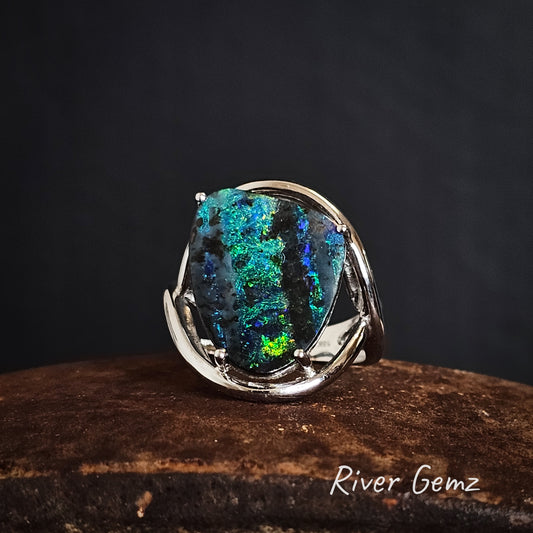 White gold ring with a large free form cut colorful boulder opal on a dark background, branded 'River Gemz'.