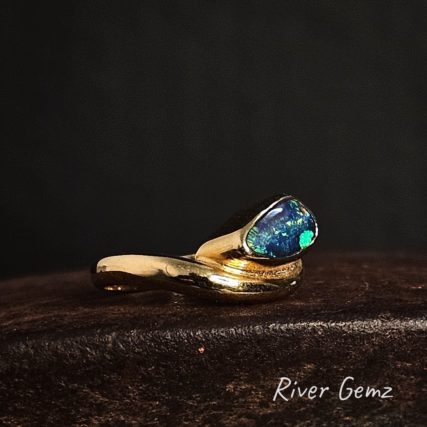 Gold ring with a blue and green opal stone on a dark background, branded 'River Gemz'.