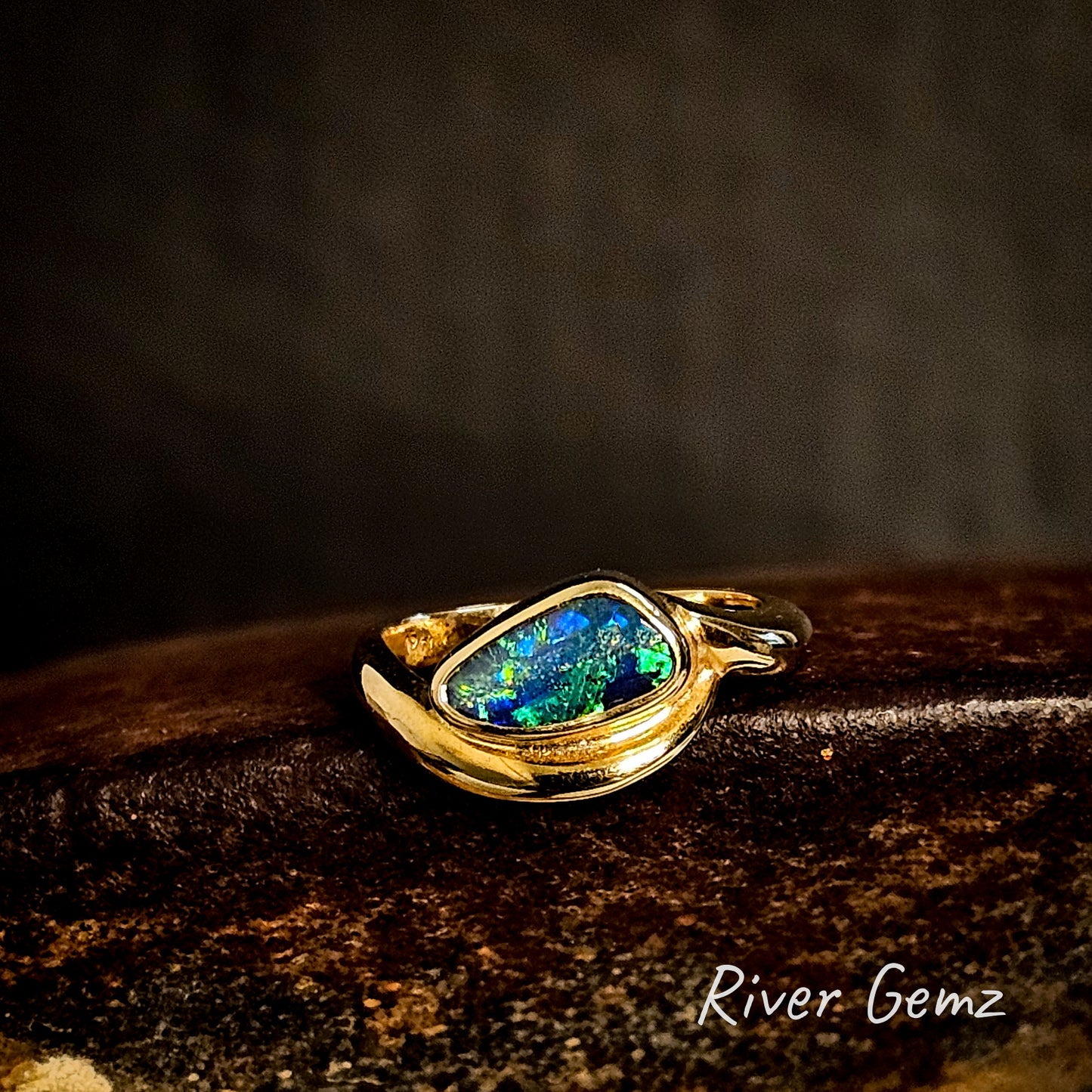 Gold ring with a free-form cut colorful boulder opal on a dark background, featuring the brand 'River Gemz'.