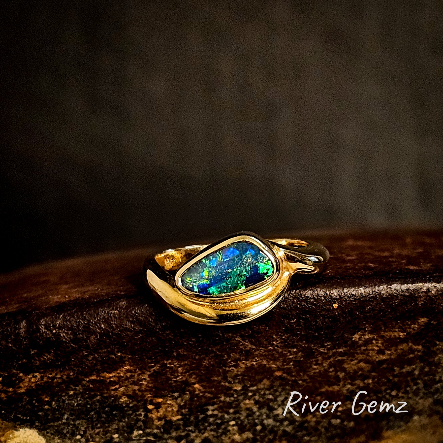 Gold ring with a free-form cut colorful boulder opal on a dark background, featuring the brand 'River Gemz'.