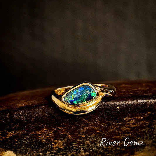 Gold ring with a free-form cut colorful boulder opal on a dark background, featuring the brand 'River Gemz'.