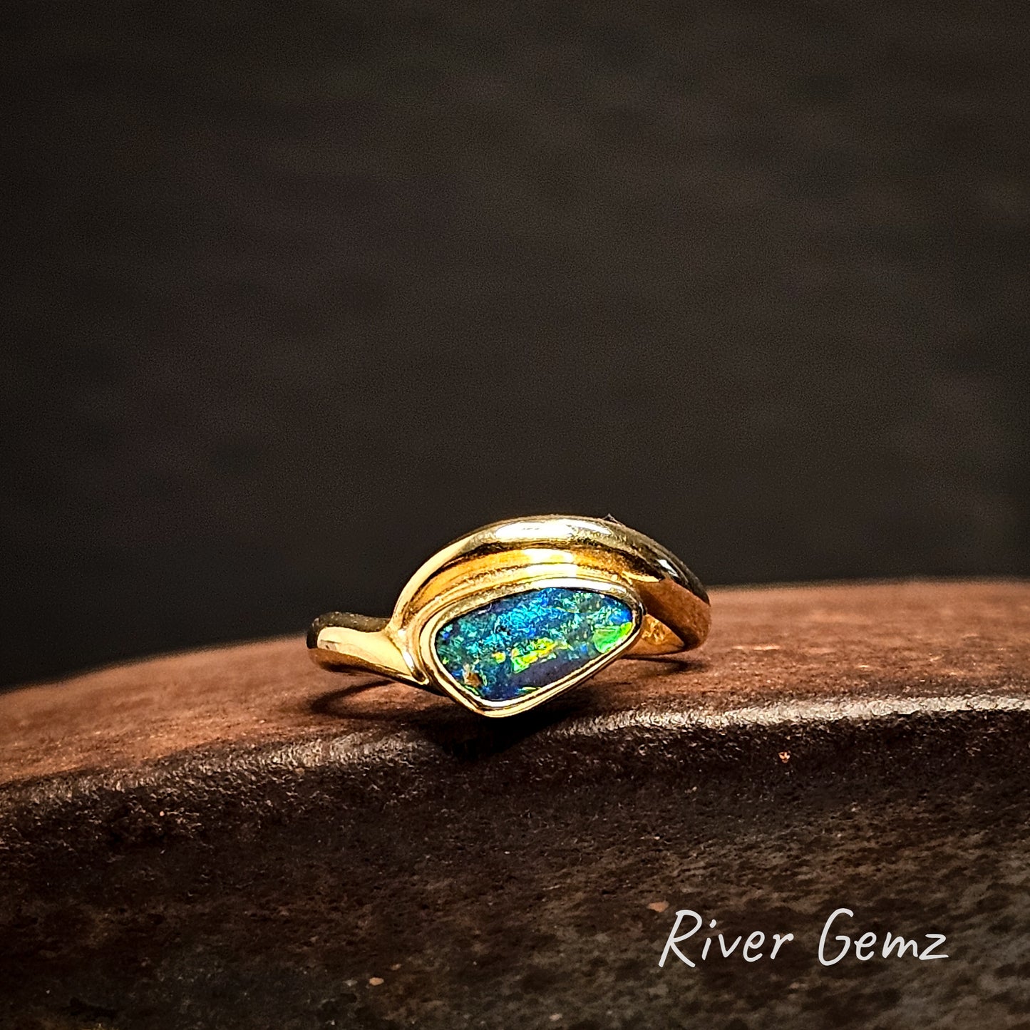 Gold ring with an opal stone on a dark surface, branded 'River Gemz'.