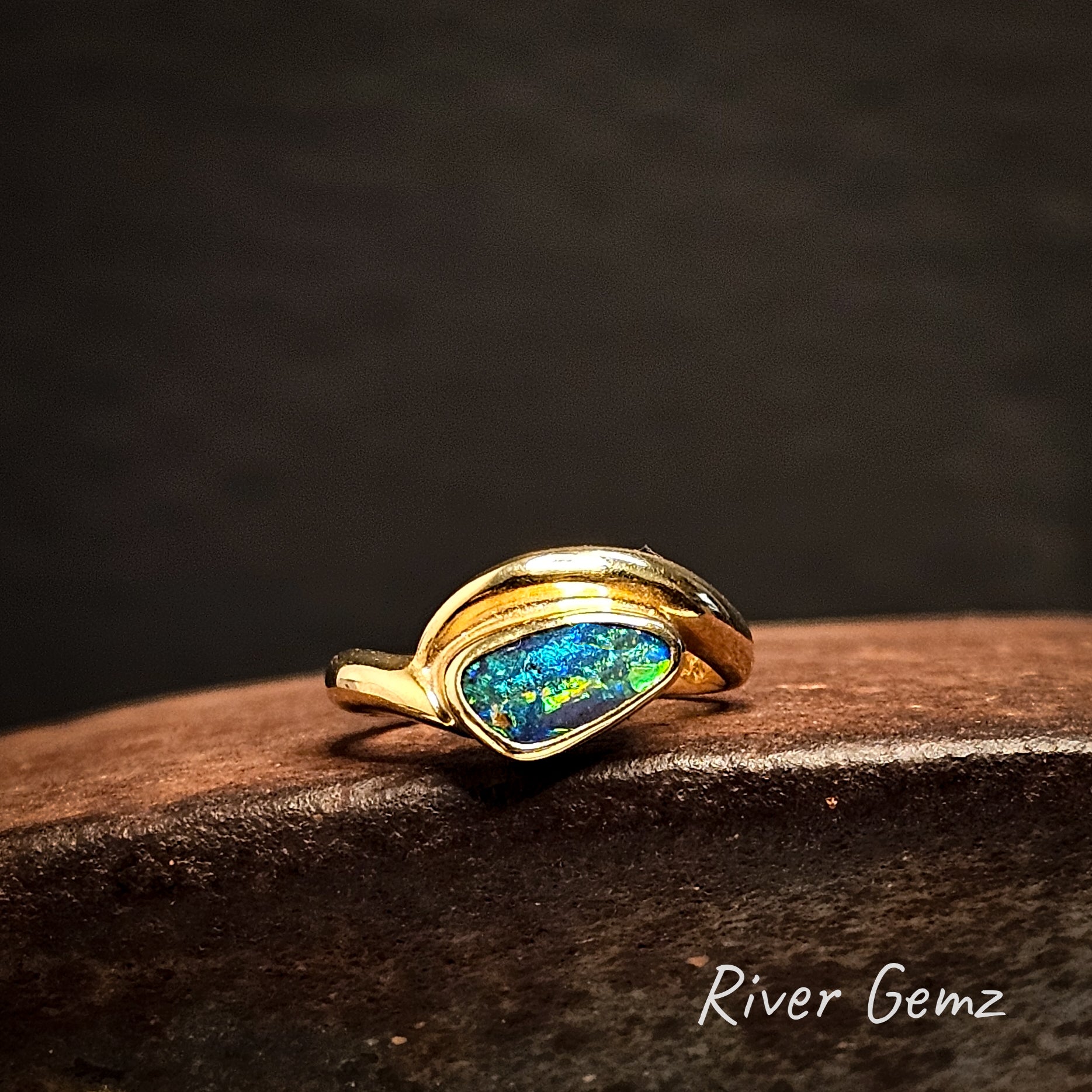 Gold ring with an opal stone on a dark surface, branded 'River Gemz'.