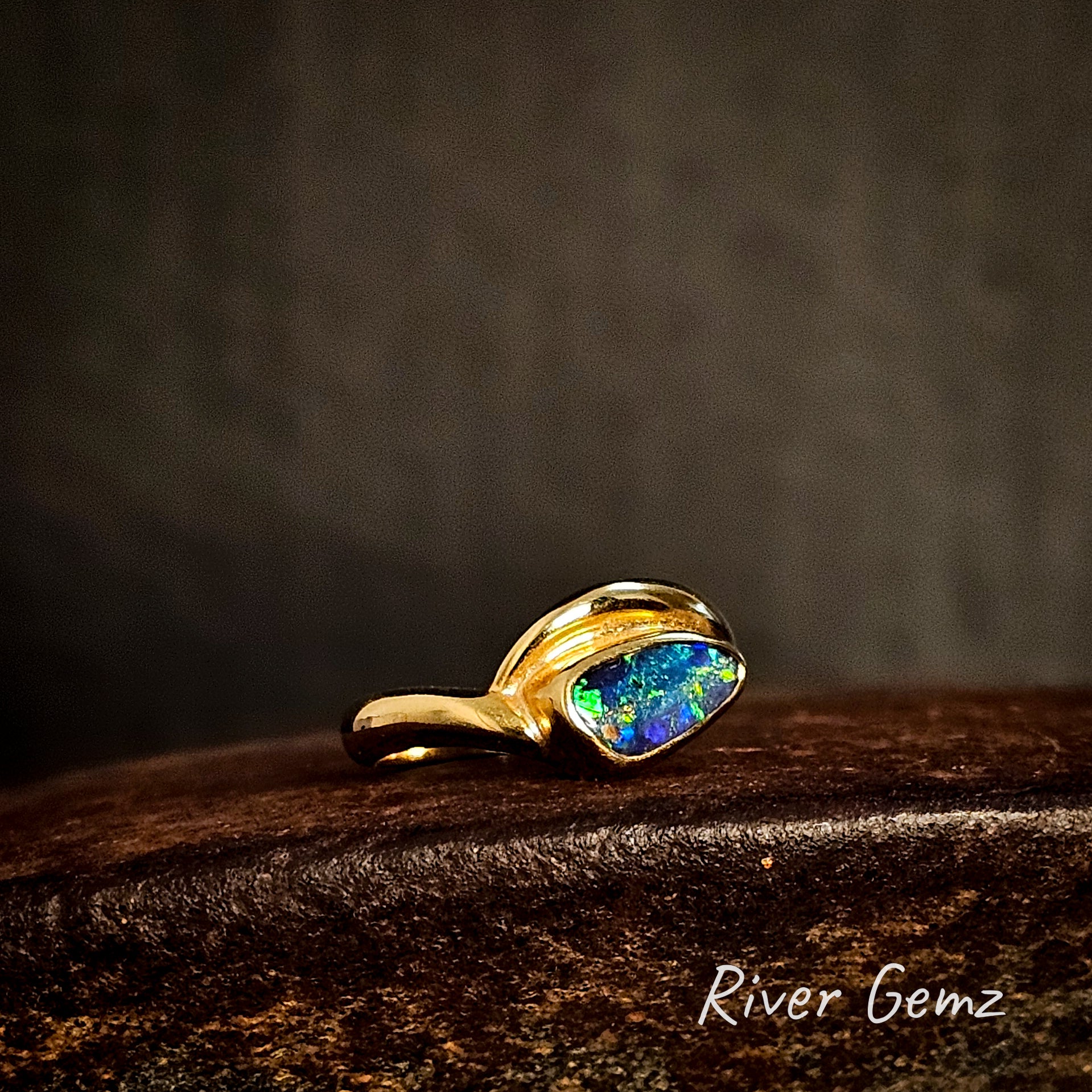 Yellow gold ring with a colorful gemstone on a dark wooden surface, branded 'River Gemz'.