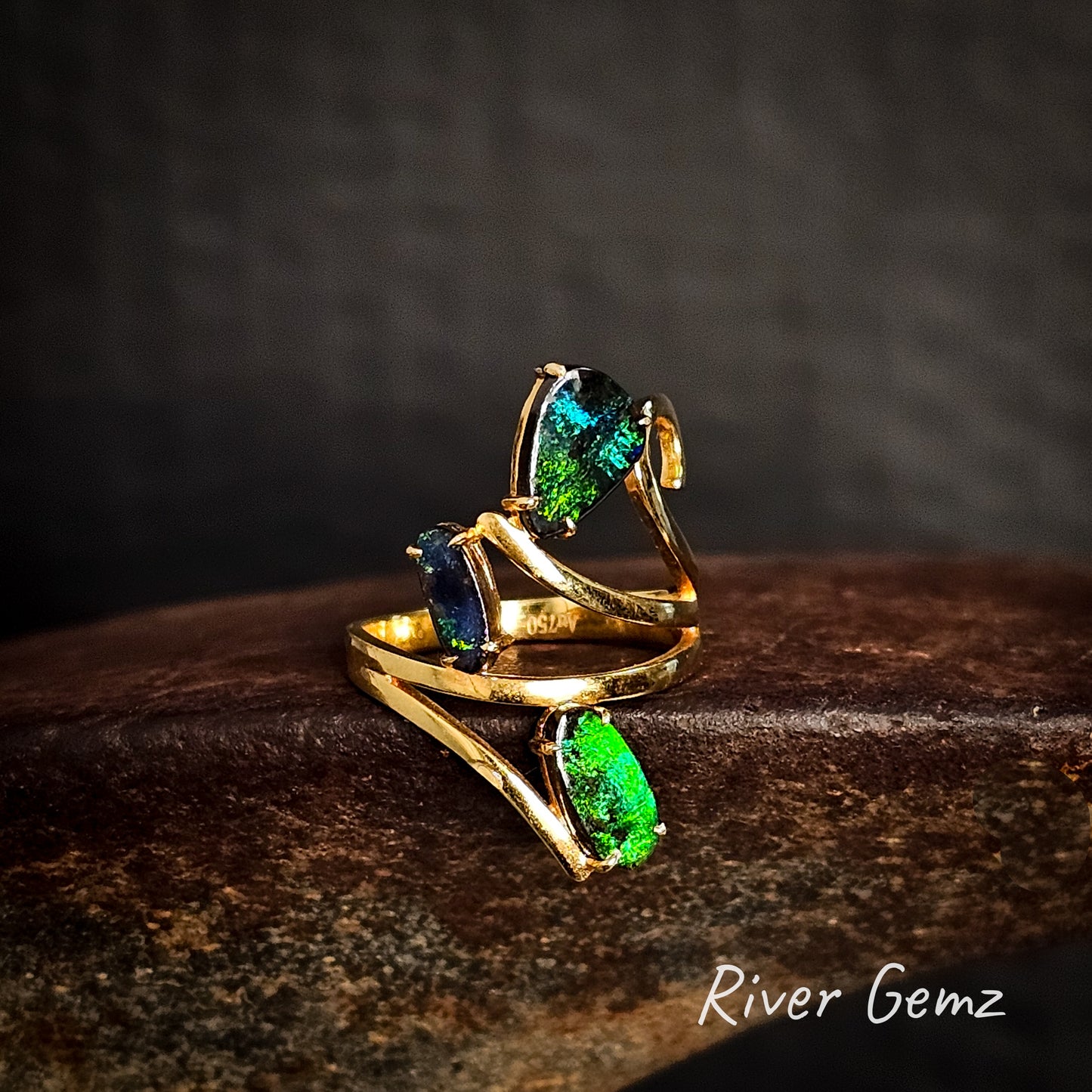 Yellow gold ring with three free-form cut green gemstones on a dark background, featuring the brand 'River Gemz'.