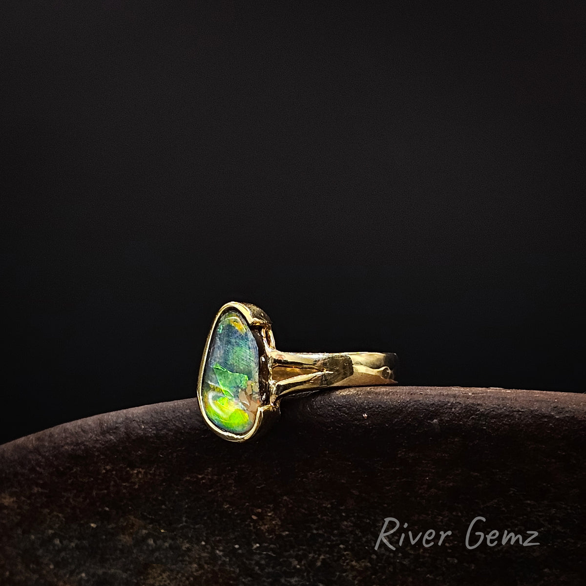 Gold ring with vibrantly coloured opal on a weathered shoe anvil against a dark background and 'River Gemz' watermark in the bottom right hand corner.