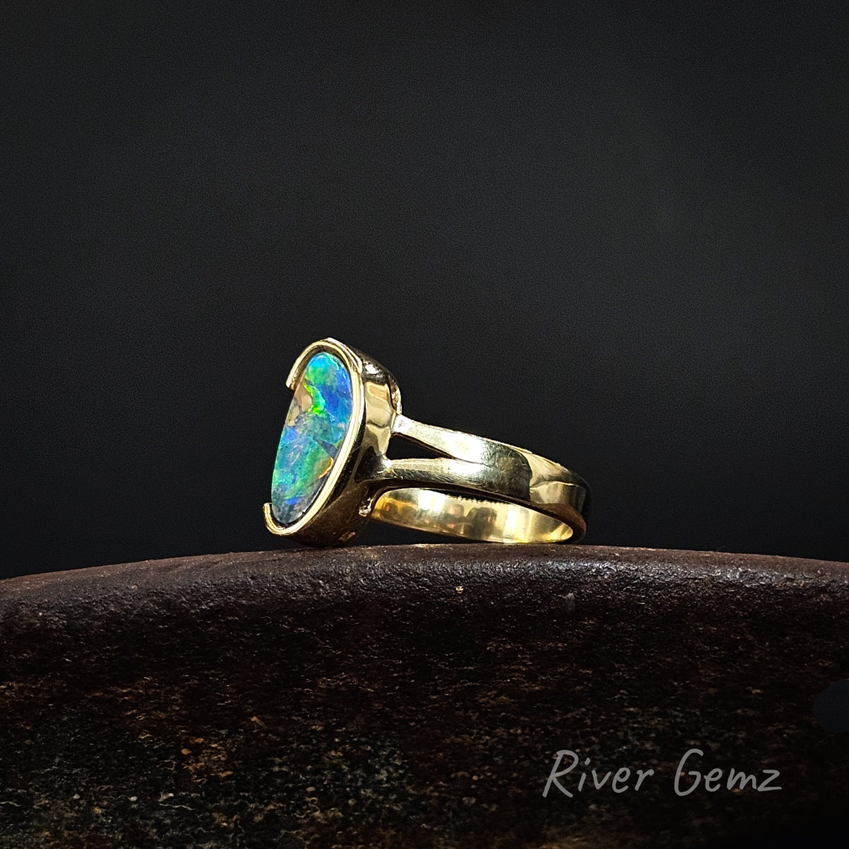 Gold ring with vibrantly coloured opal on a weathered shoe anvil against a dark background and 'River Gemz' watermark in the bottom right hand corner.