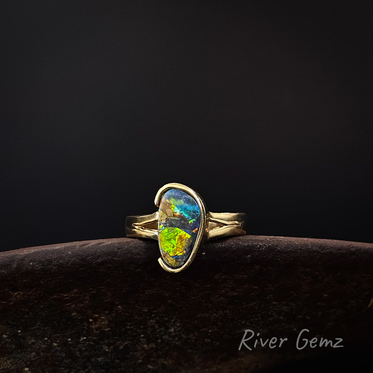Gold ring with vibrantly coloured opal on a weathered shoe anvil against a dark background and 'River Gemz' watermark in the bottom right hand corner.