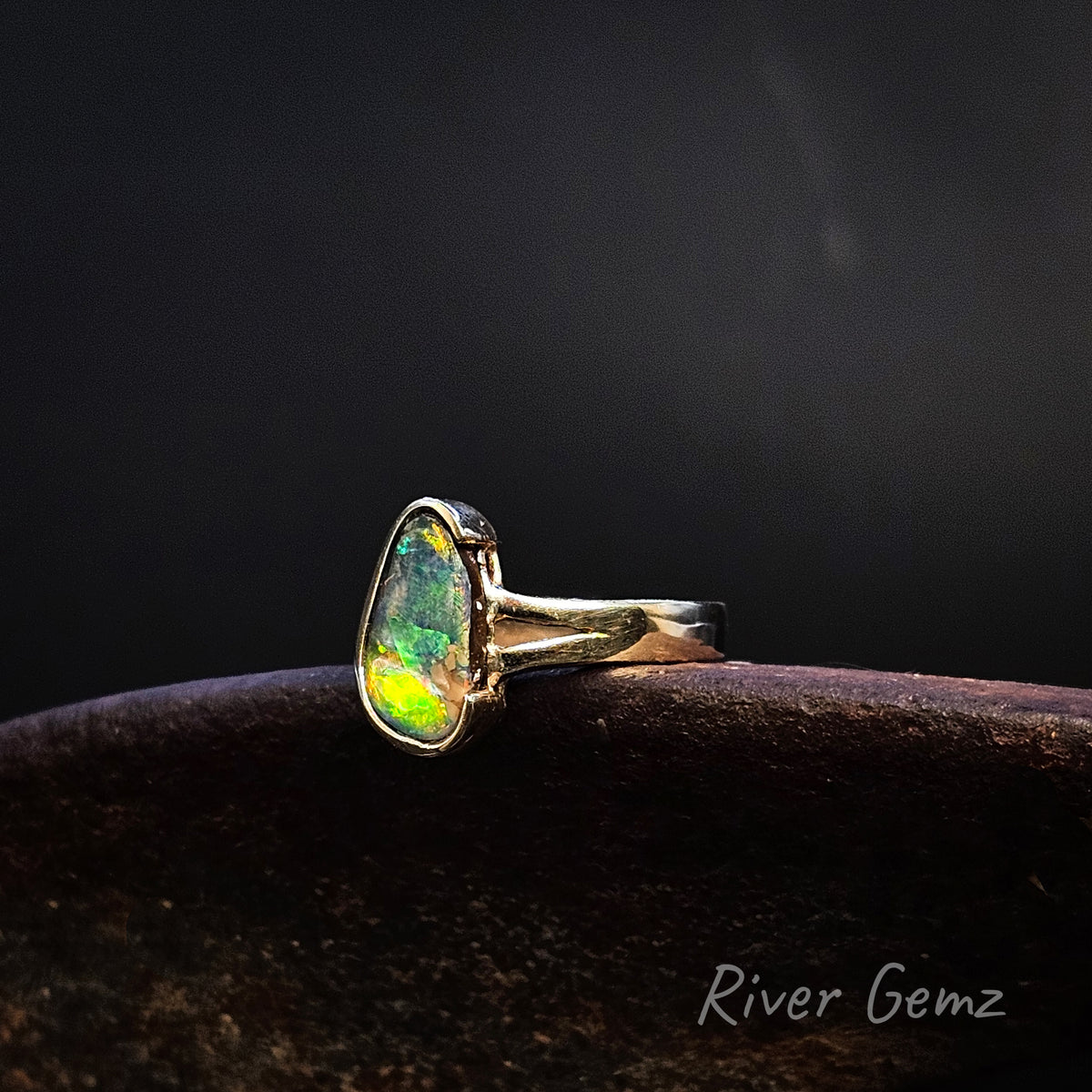 Multi-coloured free-form cut opal partial besel set in yellow gold ring against a dark background.. 'River Gemz' watermark in the bottom right hand corner.