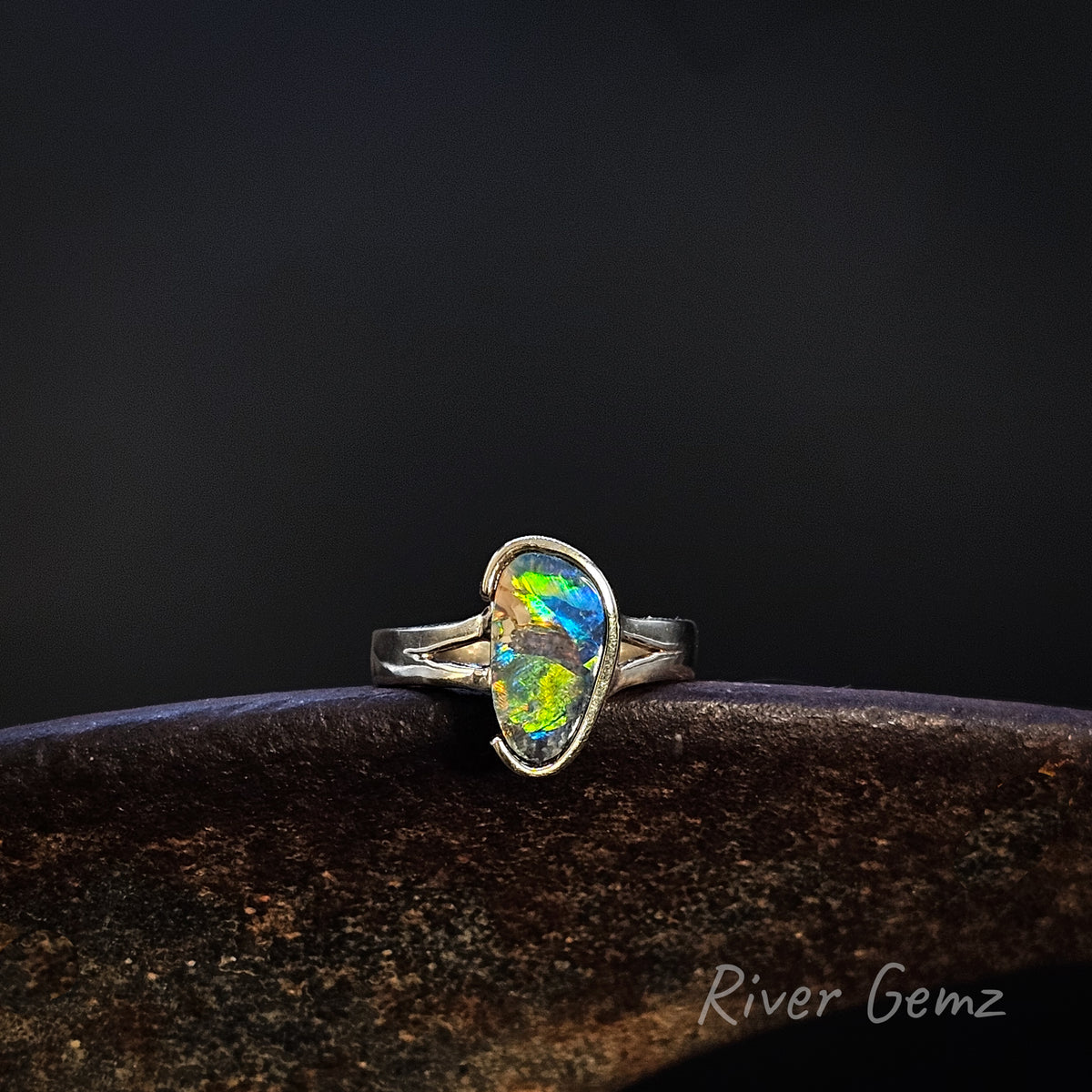 Gold ring with vibrantly coloured opal on a weathered shoe anvil against a dark background and 'River Gemz' watermark in the bottom right hand corner.