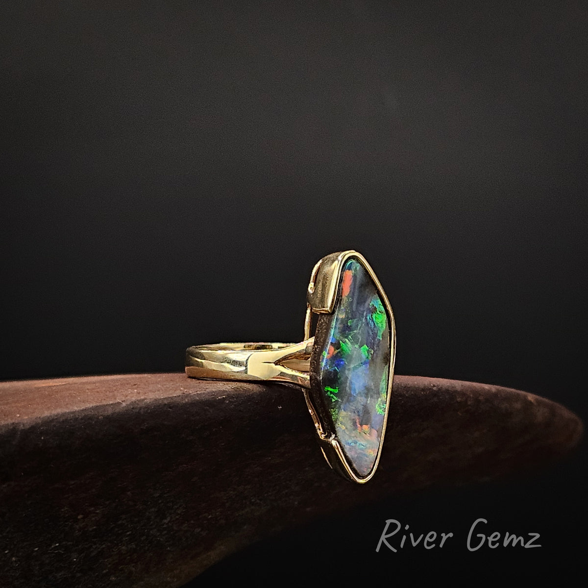 Large diamond shaped multi-coloured opal partial besel set in yellow gold ring on a rusty shoe anvil against a dark background and 'River Gemz' watermark in the bottom right hand corner.