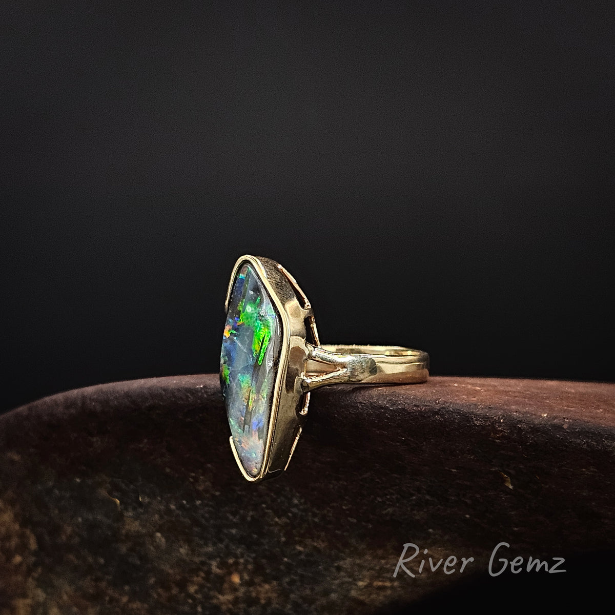 Large diamond shaped multi-coloured opal partial besel set in yellow gold ring on a rusty shoe anvil against a dark background and 'River Gemz' watermark in the bottom right hand corner.