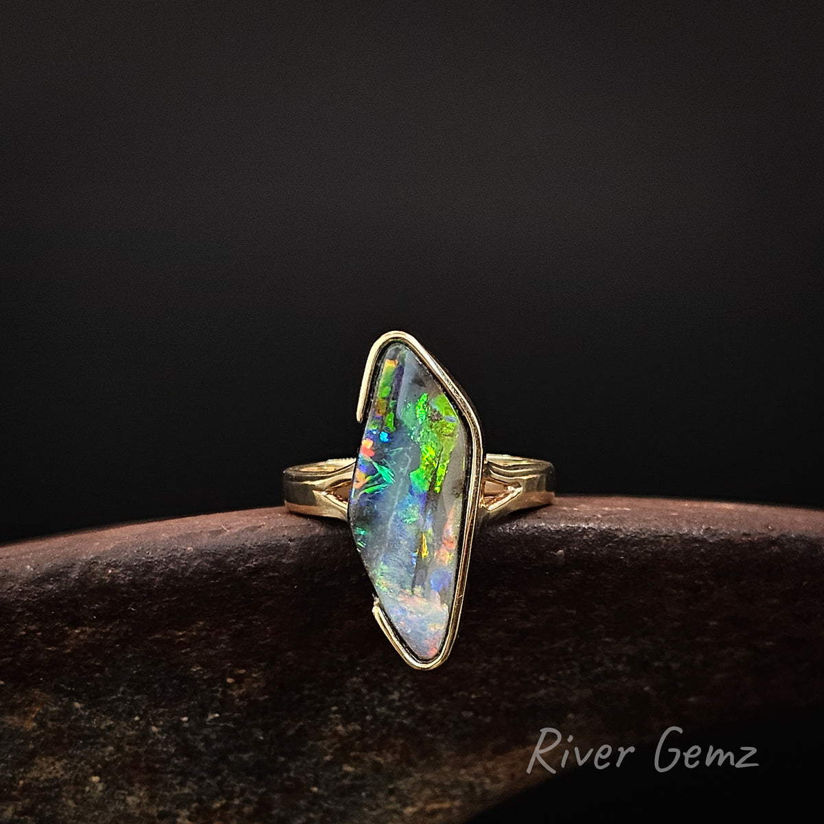 Large diamond shaped multi-coloured opal partial besel set in yellow gold ring on a rusty shoe anvil against a dark background and 'River Gemz' watermark in the bottom right hand corner.