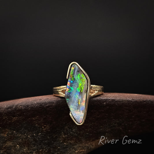 Large diamond shaped multi-coloured opal partial besel set in yellow gold ring on a rusty shoe anvil against a dark background and 'River Gemz' watermark in the bottom right hand corner.