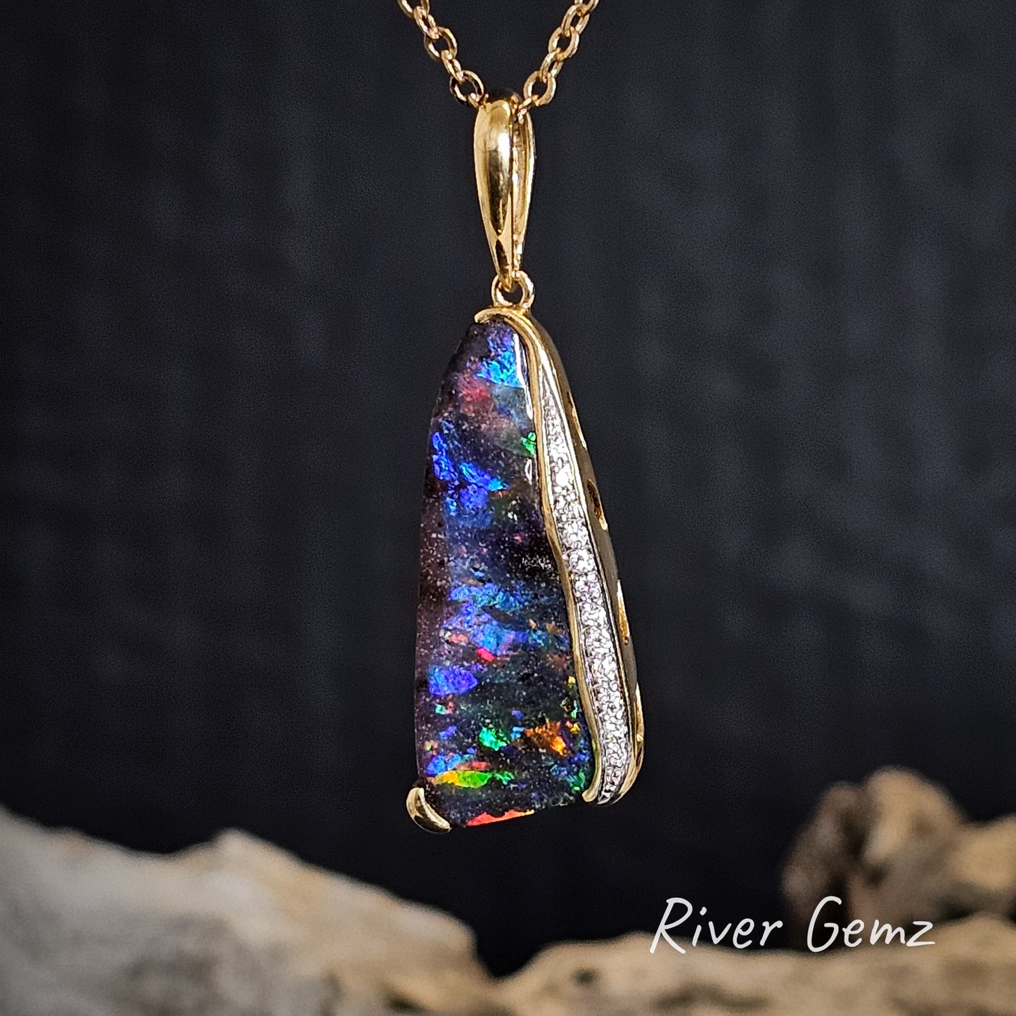 Multi-coloured dark opal and diamonds pendant with gold setting on a dark background, featuring the brand 'River Gemz'.