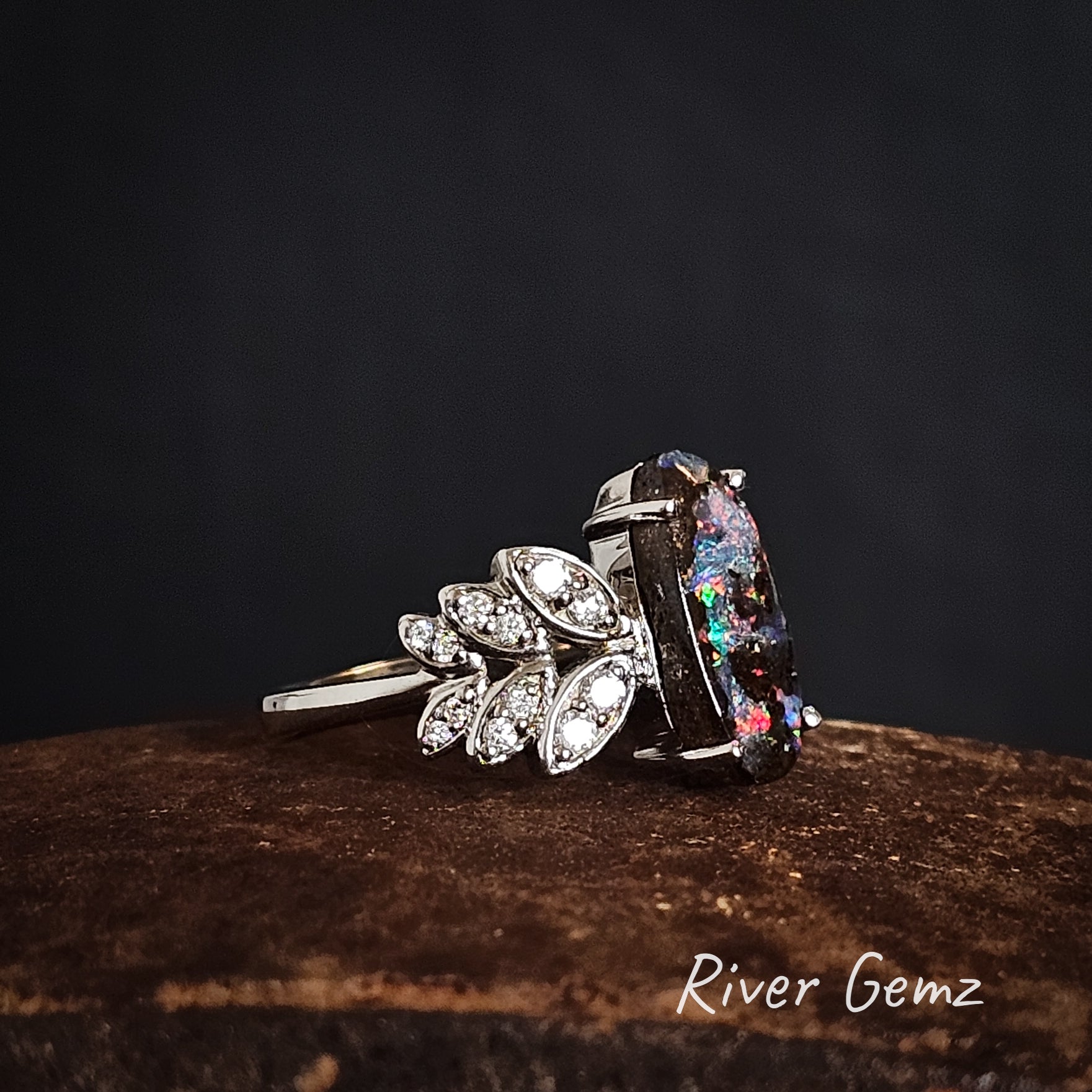 Side view of the white gold ring with multi-coloured boulder opal and diamond accents in 6 leaves on the shoulders, on a dark background with 'River Gemz' branding.
