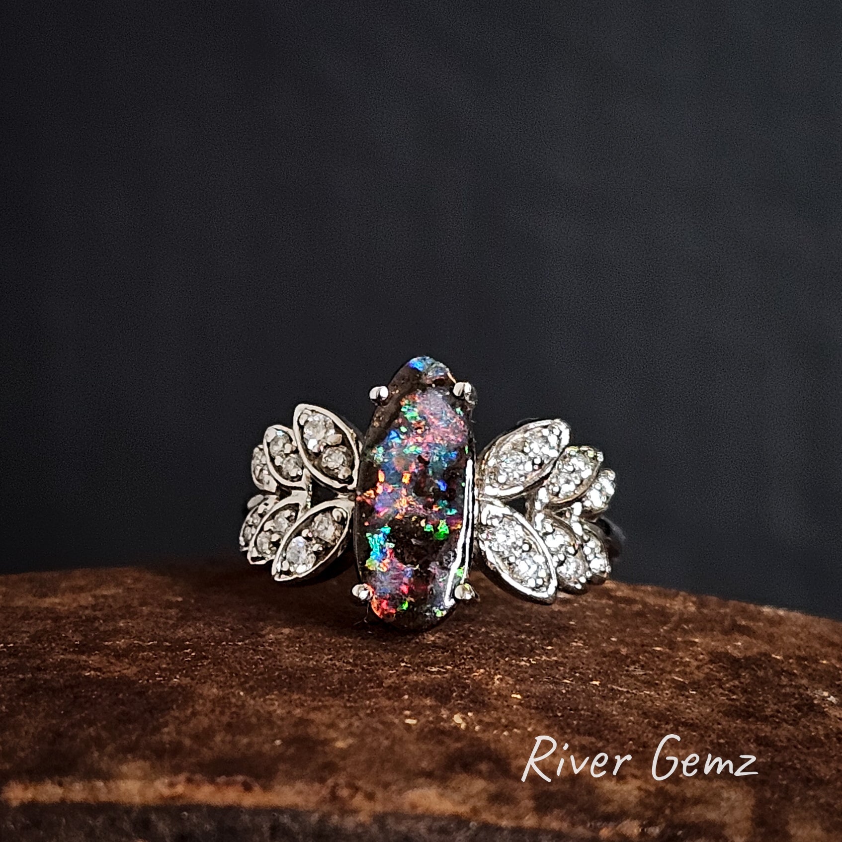 Colourful opal claw set in white gold ring with diamond leaves on the shoulders on a dark background, branded 'River Gemz'.