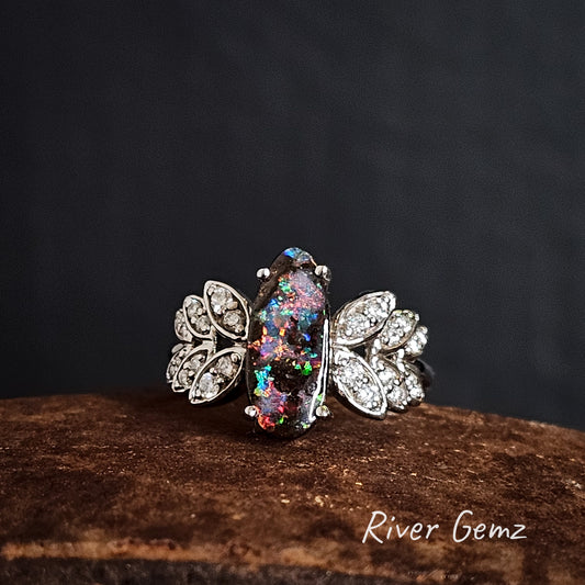 Colourful opal claw set in white gold ring with diamond leaves on the shoulders on a dark background, branded 'River Gemz'.