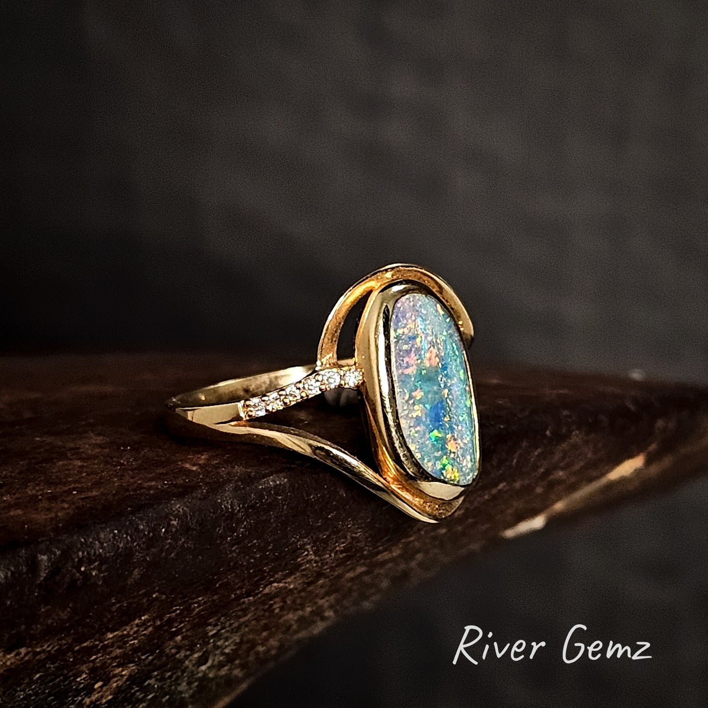 Gold ring with an opal stone and diamonds on the shoulders on a dark surface, branded 'River Gemz'.