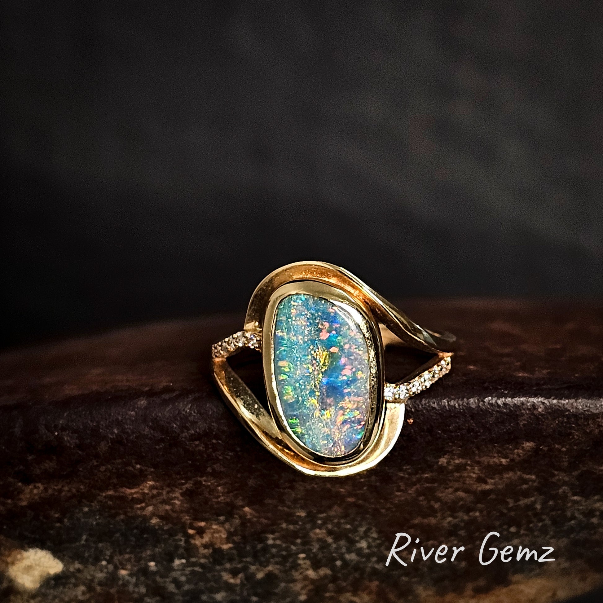 Gold ring with a large colorful boulder opal and diamonds on a dark background, featuring the brand 'River Gemz'.